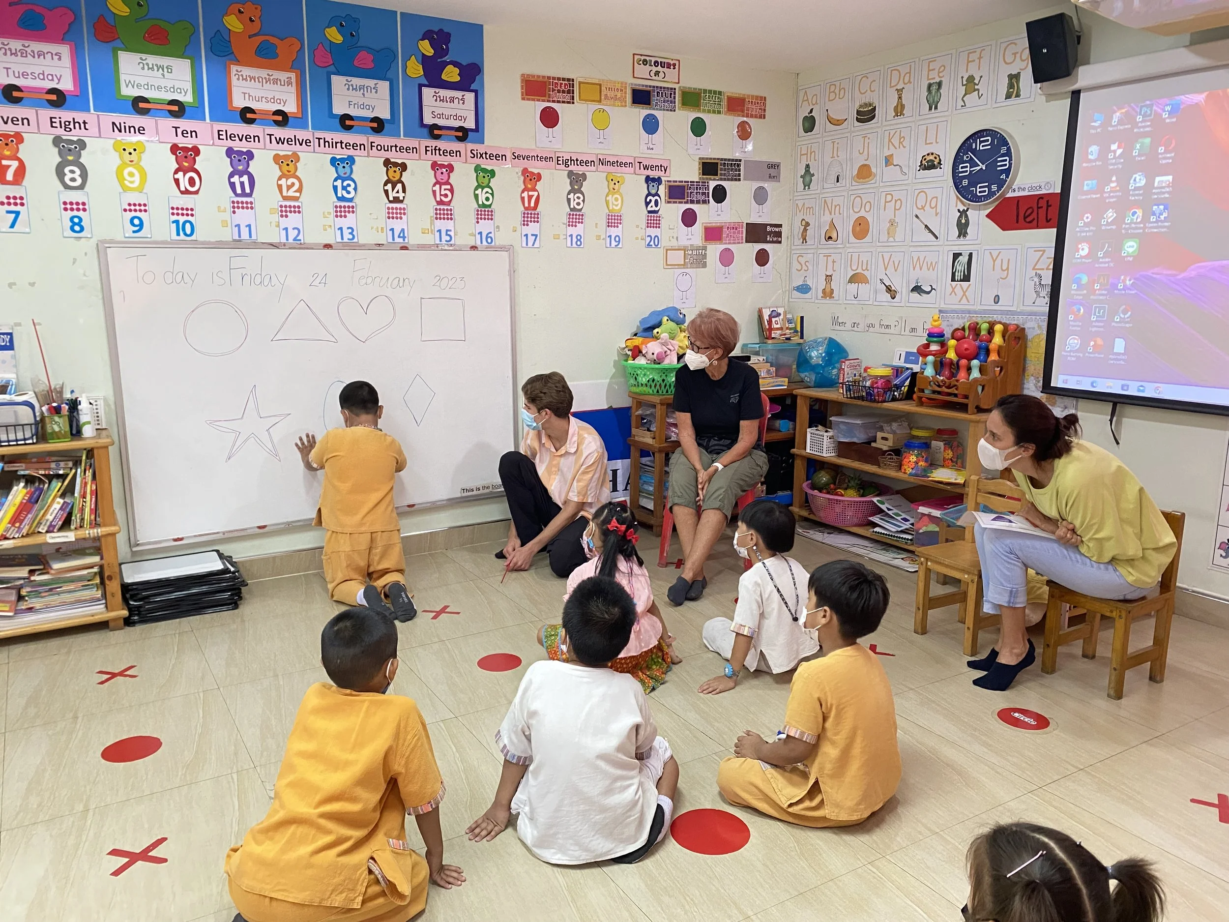 In a classroom, children are sitting on the floor facing a whiteboard where a teacher is drawing shapes. A boy is standing and drawing on the board. Two teachers are sitting, supervising the children, all wearing masks. The classroom has colorful educational decorations, including numbers, days of the week, and alphabet posters.