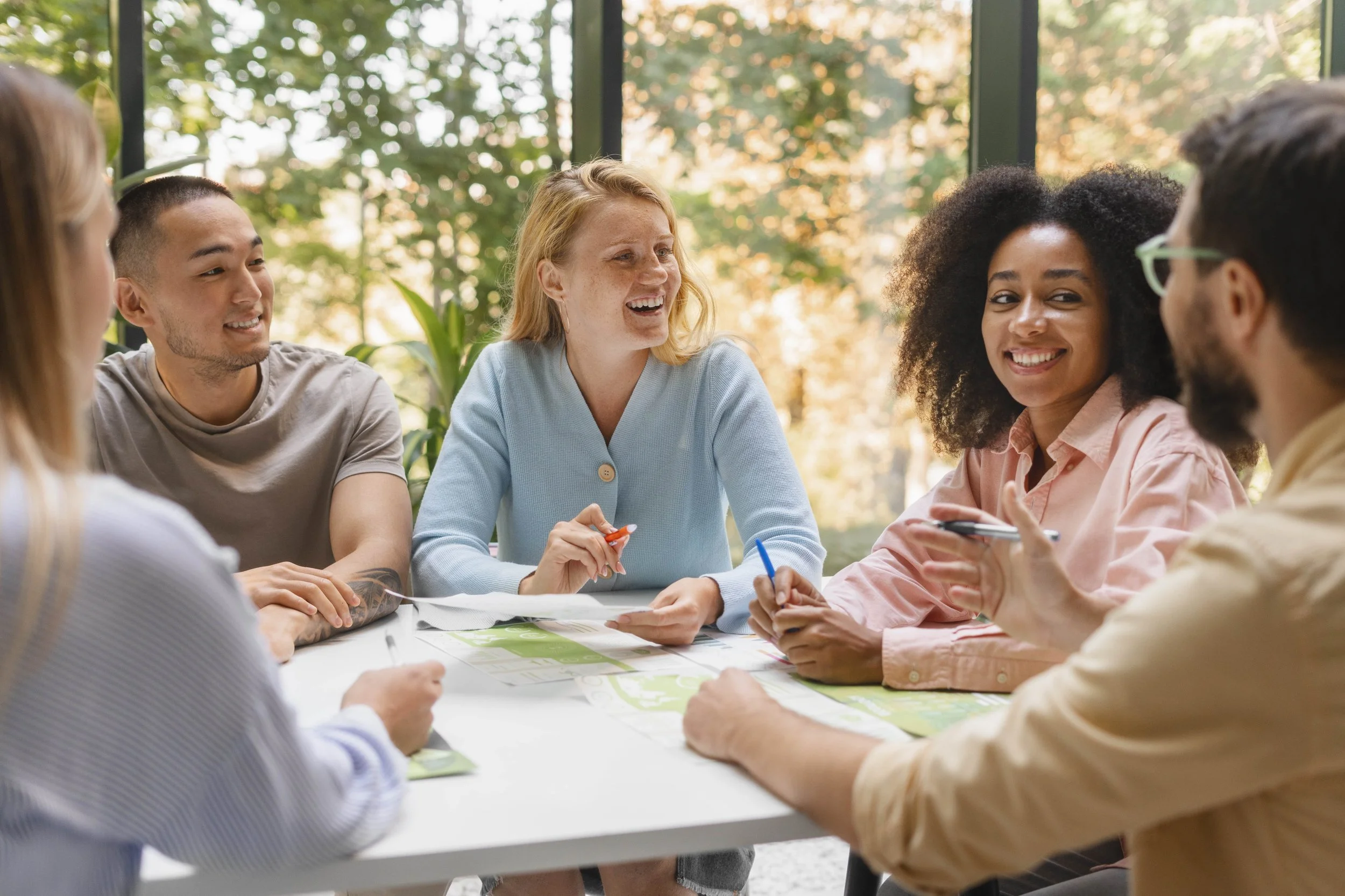Group of six diverse people sitting around a table having a discussion in a bright, glass-walled room with trees outside.