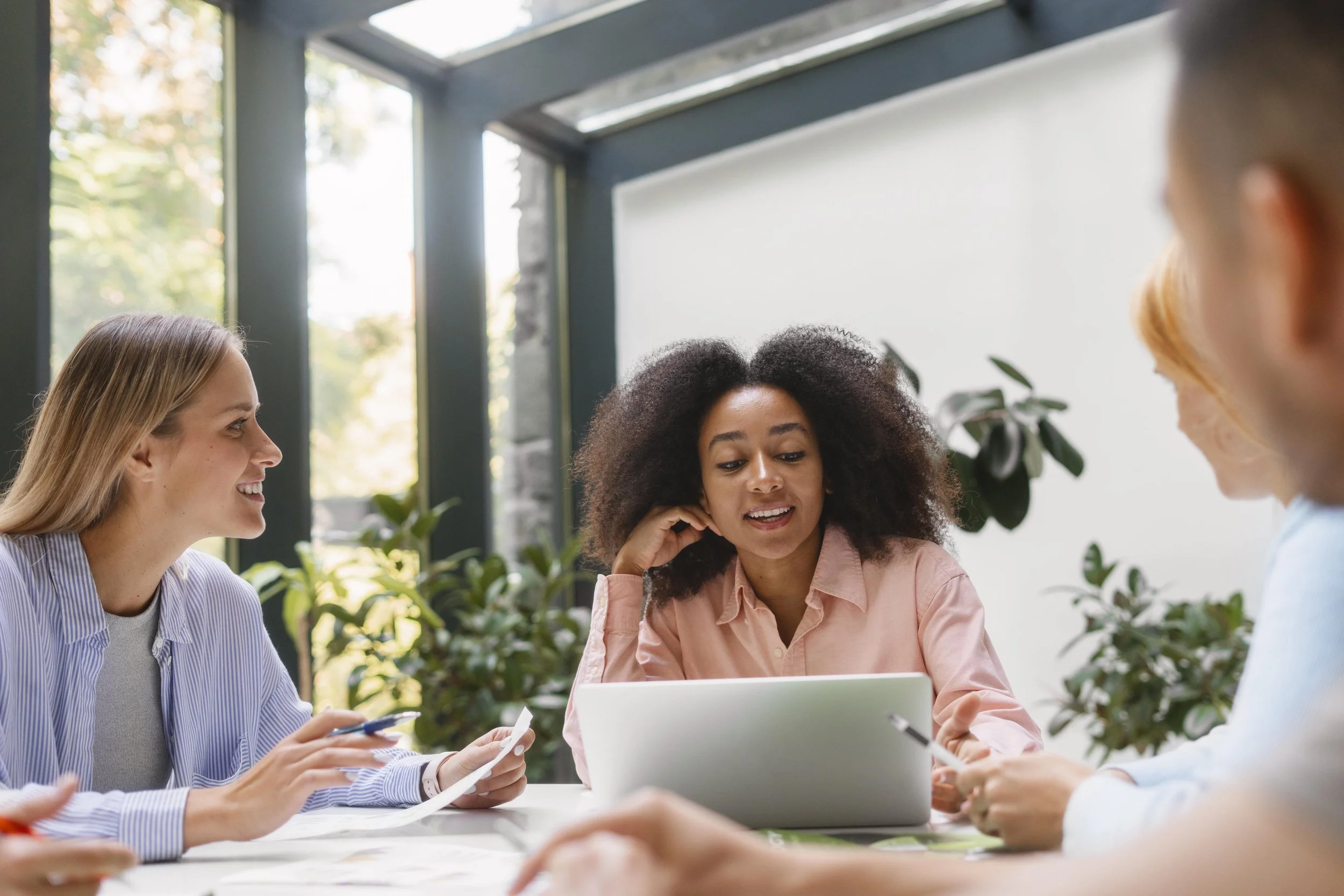Group of four women in a meeting room having a discussion, with one woman looking at a laptop.