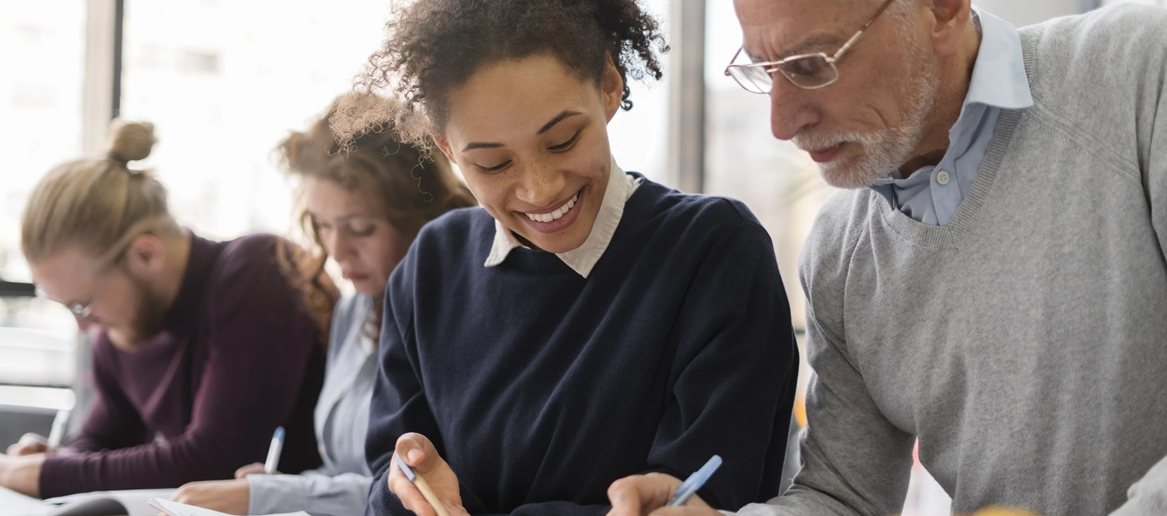 A diverse group of people working together at a table, smiling and writing notes, in a bright and modern office or classroom setting.