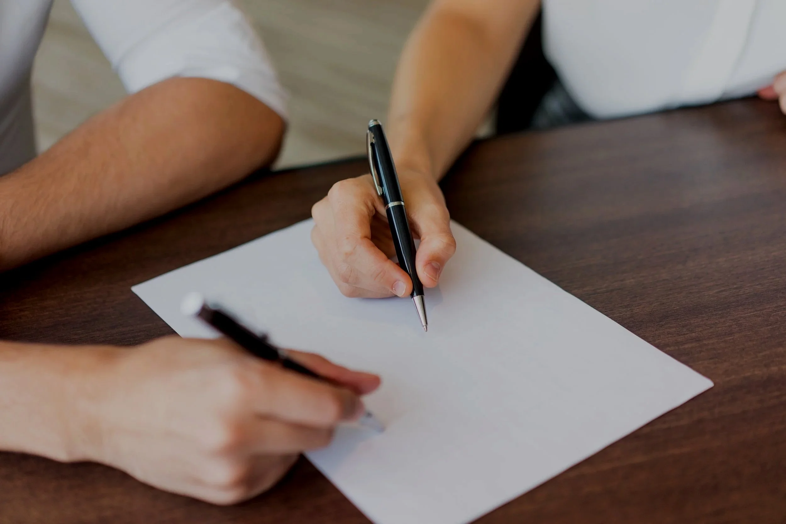Two people sitting at a wooden table, one with a pen and the other holding a paper, engaged in writing or signing.