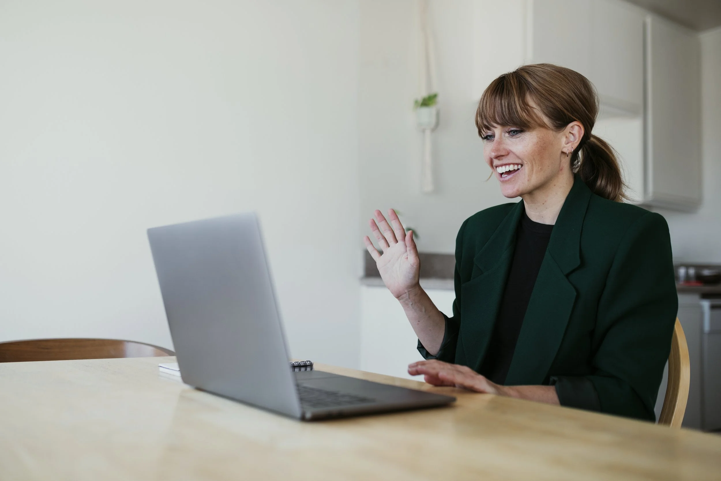 A woman with brown hair, wearing a dark green blazer, sitting at a wooden table, smiling and waving while using a silver laptop in a bright room.