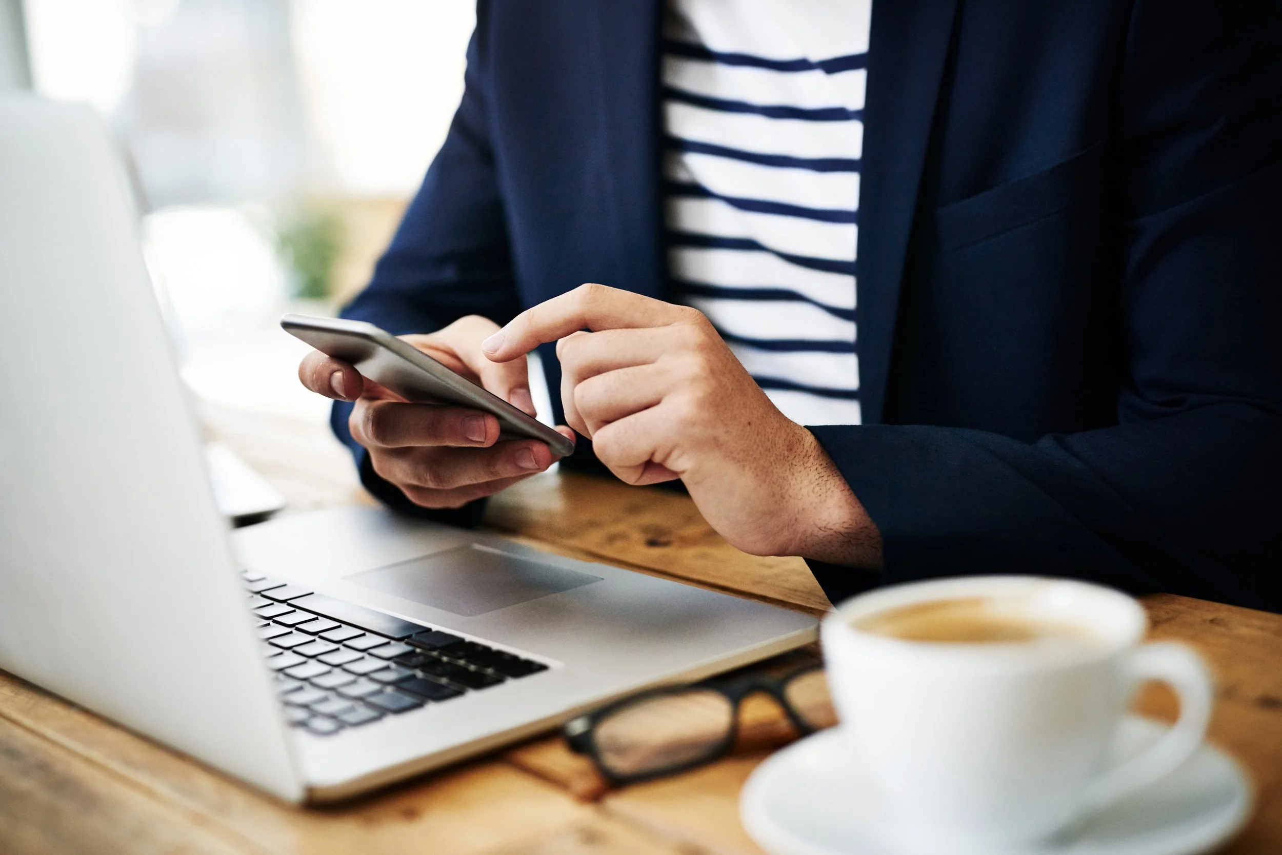 Person in a navy blazer and striped shirt using a smartphone at a wooden table with a laptop, glasses, and a cup of coffee.