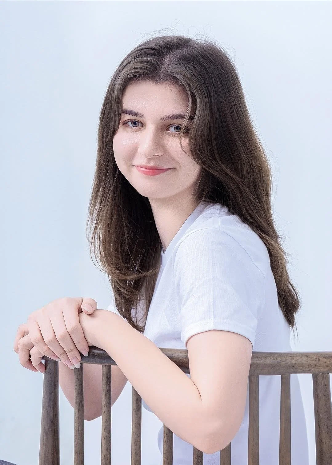 A young woman with long brown hair and wearing a white t-shirt is smiling and resting her arms on a wooden chair, with a light blue background.