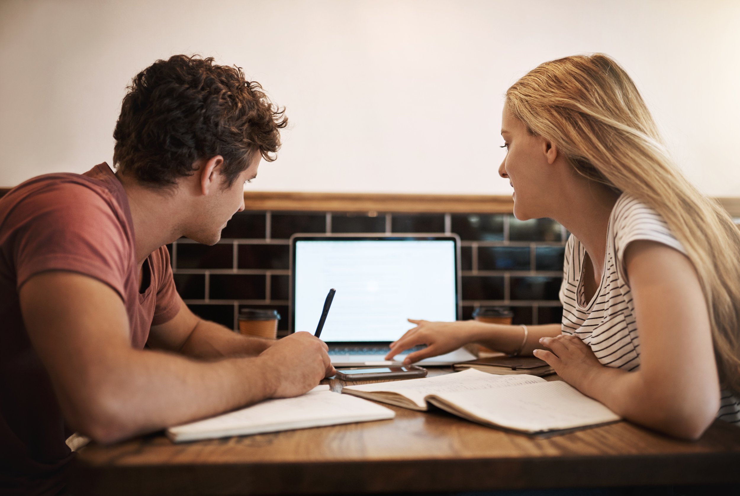 A man and woman sitting at a table working together with notebooks, a laptop, and coffee cups in a cafe.