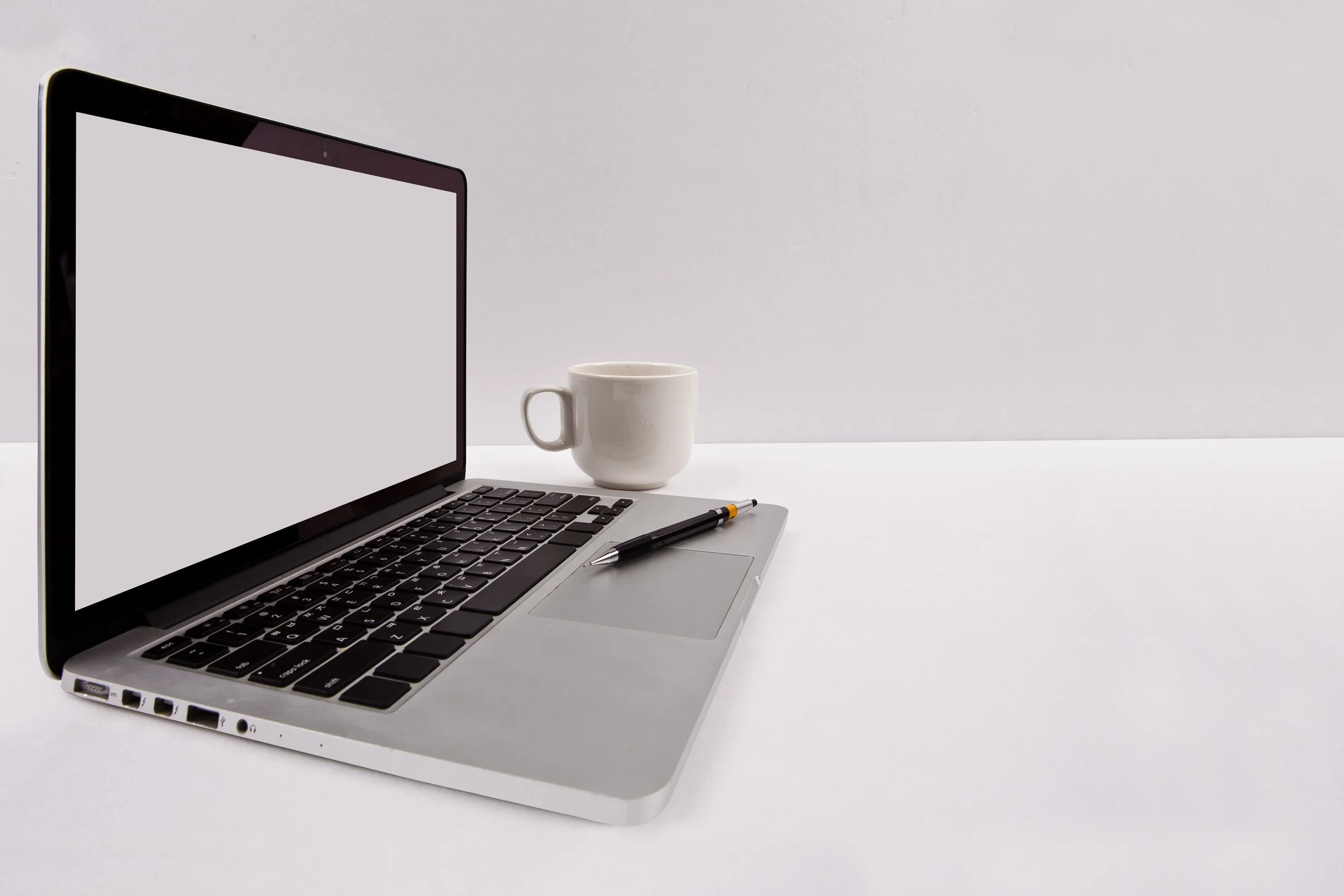 Open silver laptop with black keyboard on a white table, white coffee mug, and black pen next to the laptop.