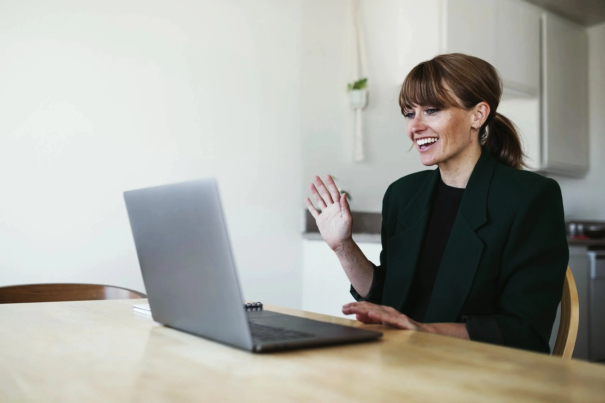 A woman with brown hair, wearing a black jacket, smiling and waving while sitting in front of a laptop at a wooden table in a kitchen.
