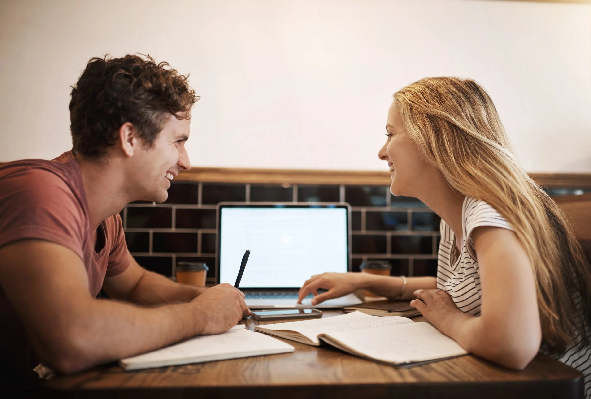 A young man and woman smiling and laughing during a meeting or study session at a table with open books, a laptop, and a smartphone.