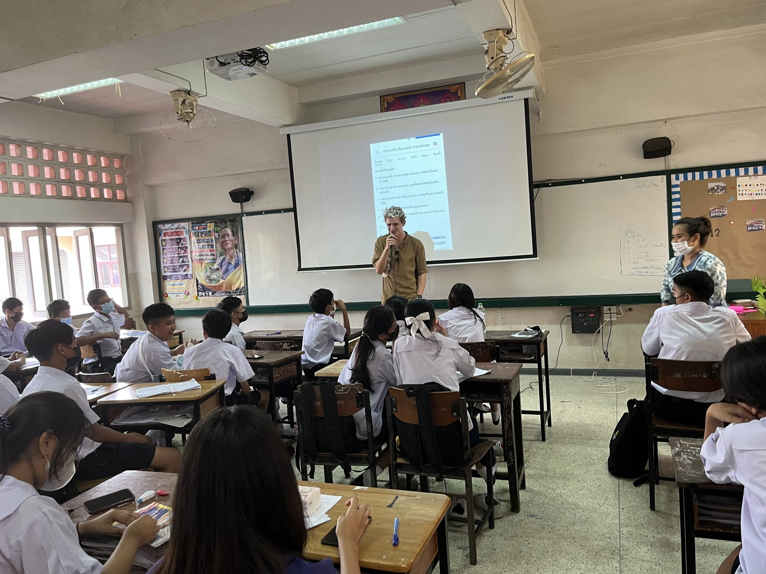 A classroom with students wearing white uniforms and face masks, sitting at desks, listening to a teacher standing in front of a projector screen. The teacher, a blond man, is speaking into a microphone. Another woman, also wearing a face mask, stands nearby. The classroom has a whiteboard, posters, and windows on the left side.