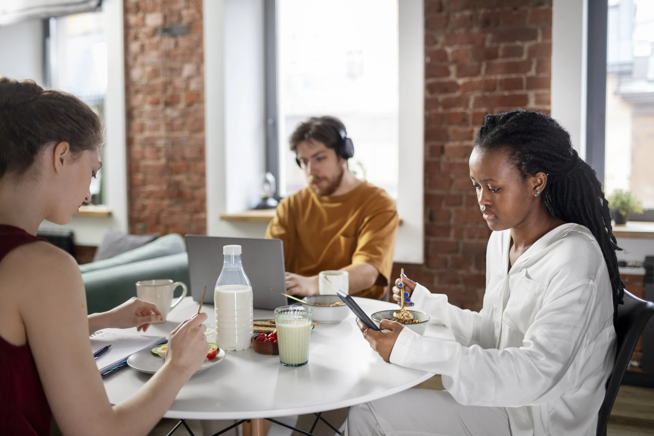 Three young adults sitting at a white table in a bright, modern room with brick walls, eating breakfast and using electronic devices.