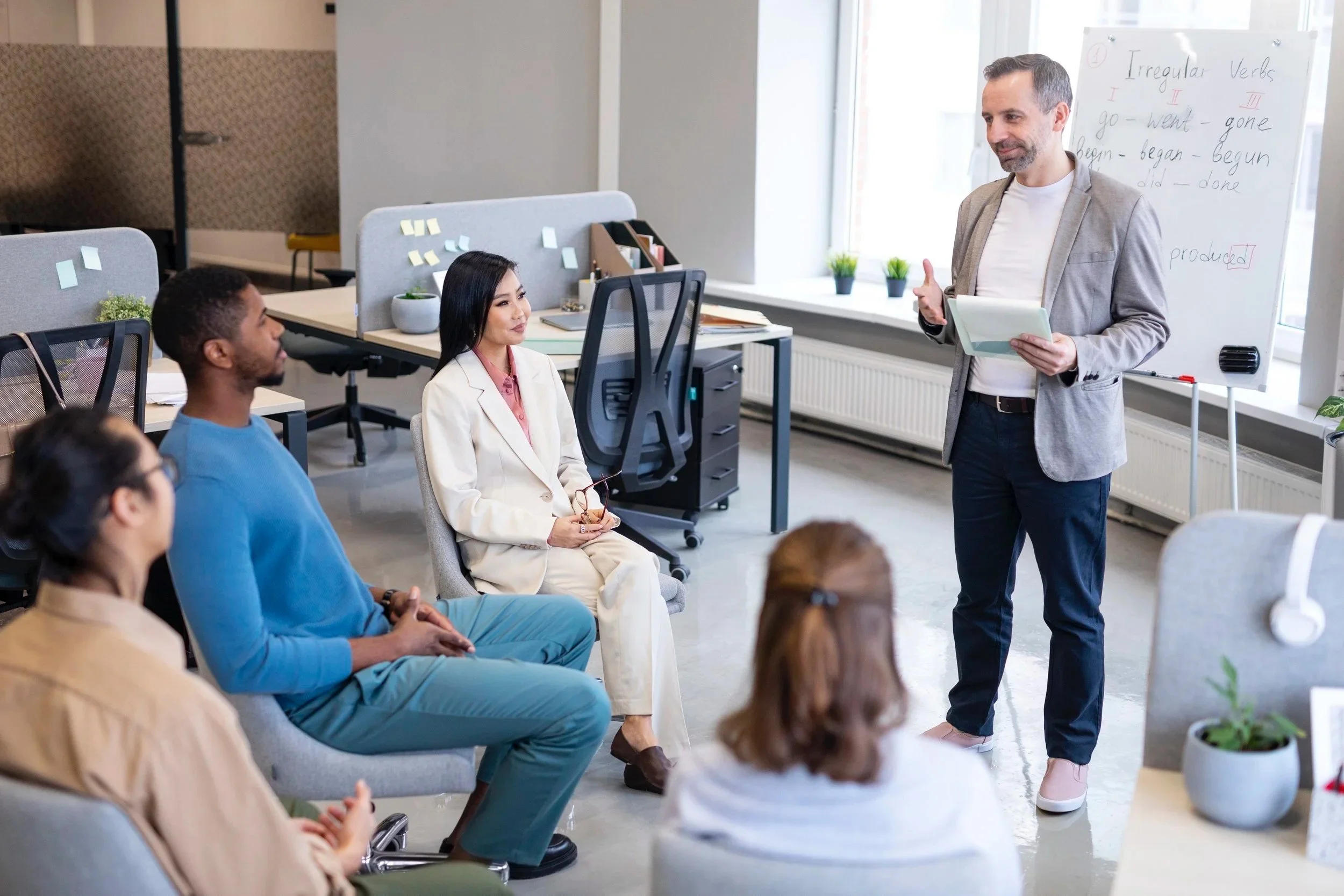 A man giving a presentation to a group of five seated diverse adults in a modern office space with a whiteboard.