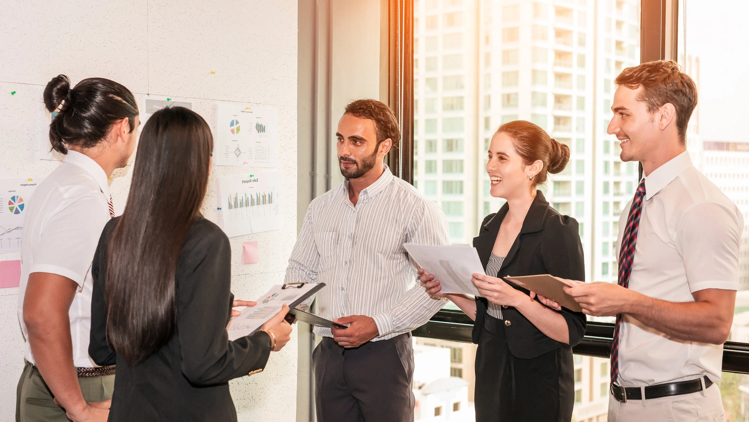 Five business professionals standing in a conference room near a window, discussing and holding documents, with charts on the wall.