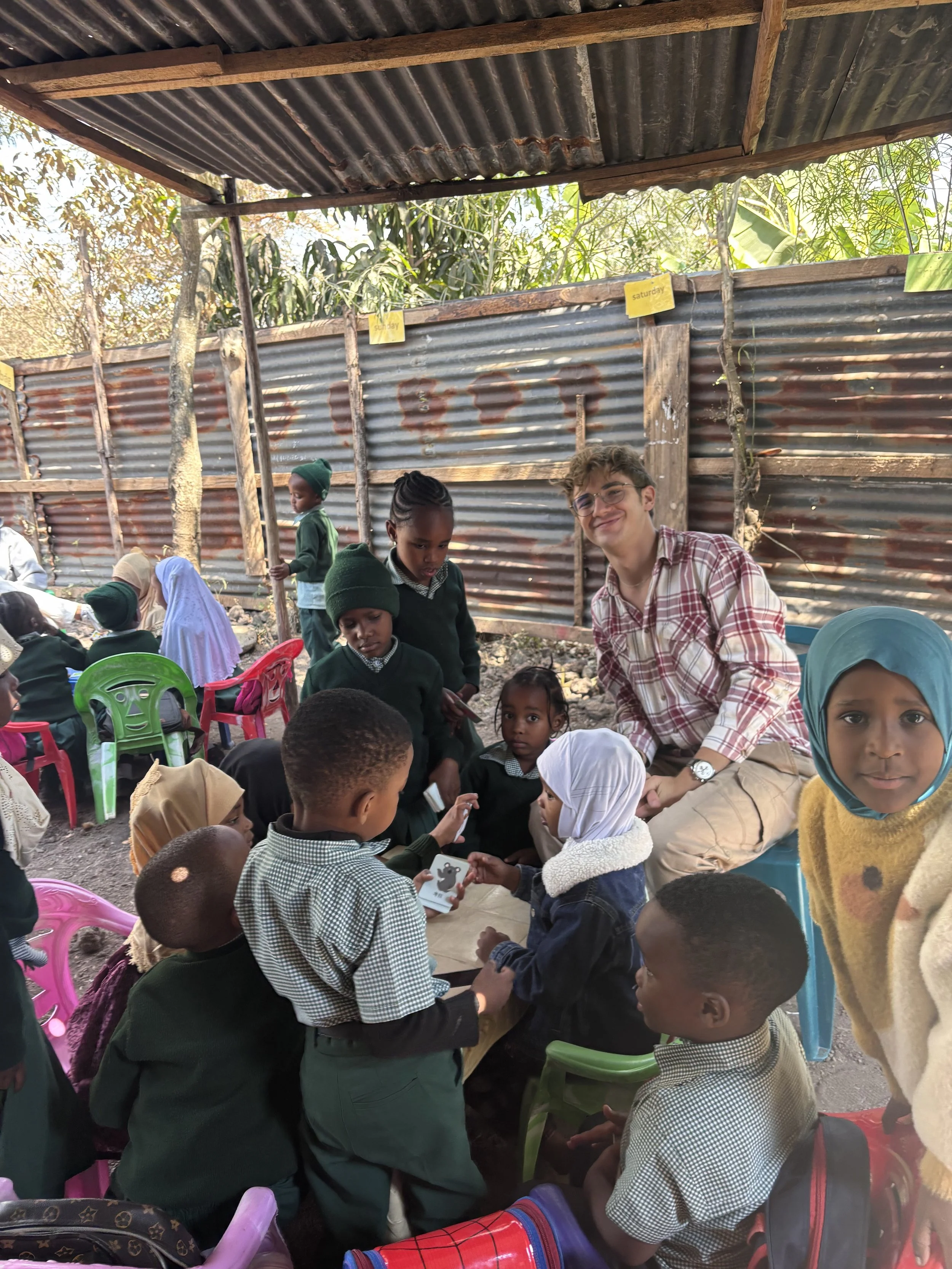 Children and a young man gathering outdoors under a corrugated metal and wood shelter, with some holding cards and others sitting on colorful plastic chairs, surrounded by a rustic fence and trees.