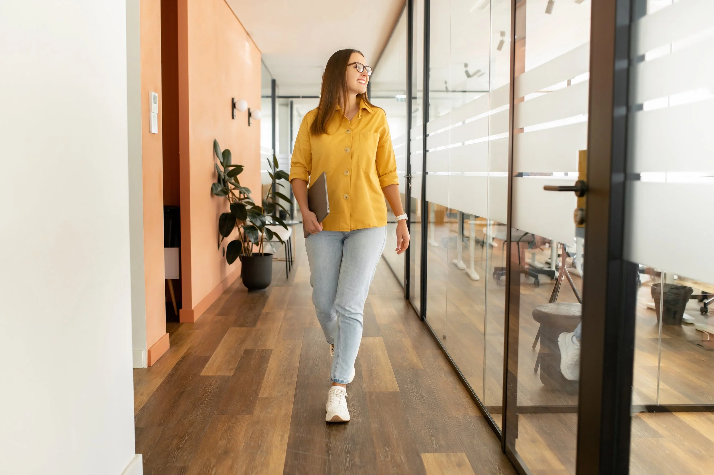 A young woman with glasses, wearing a yellow shirt and light jeans, walking down an office hallway while holding a laptop.