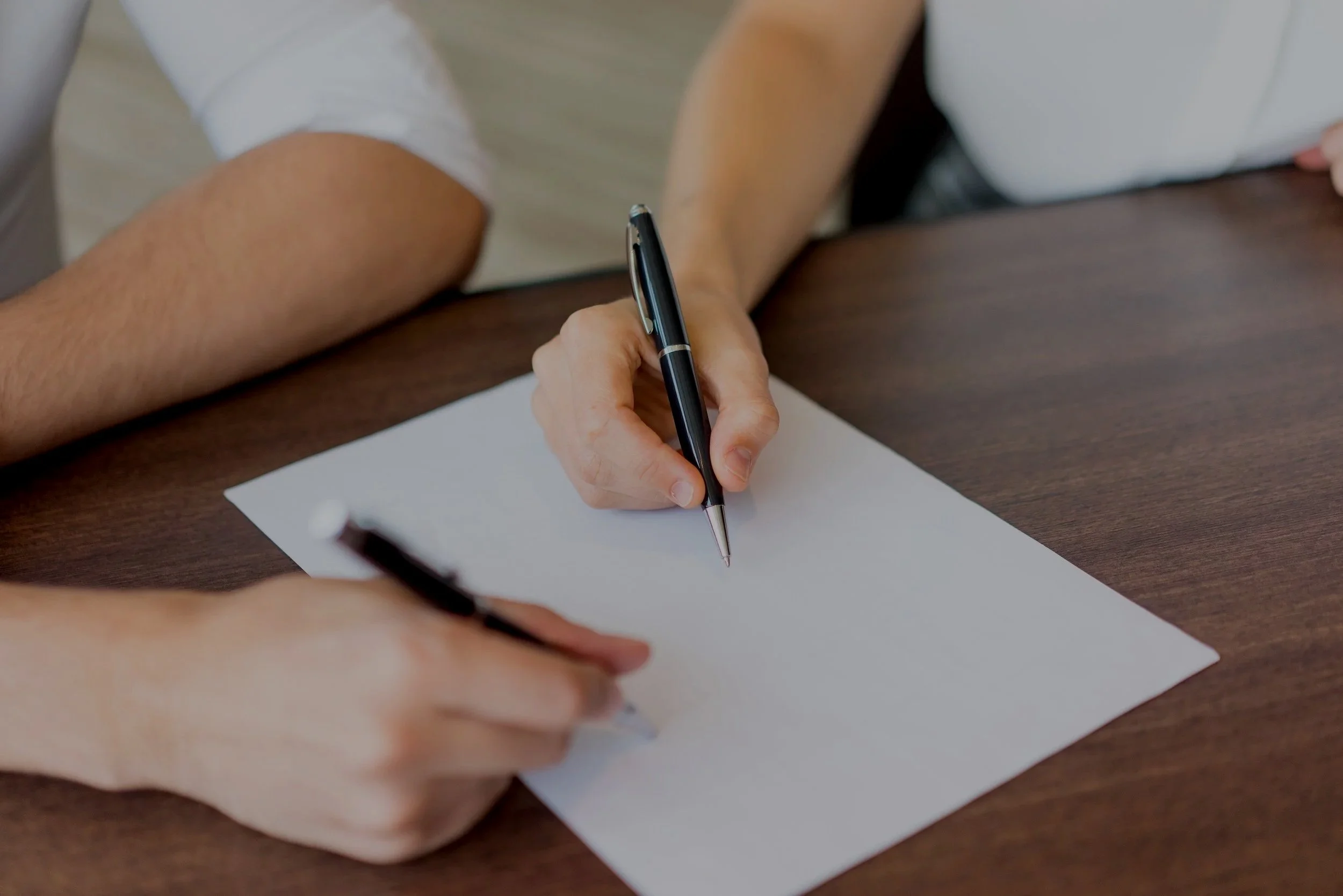 Two people are sitting at a wooden table, writing on blank white paper with black pens.