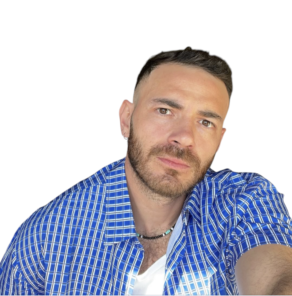 A man with short dark hair, beard, and earrings, wearing a blue checkered shirt and a necklace, taking a selfie against a white background.