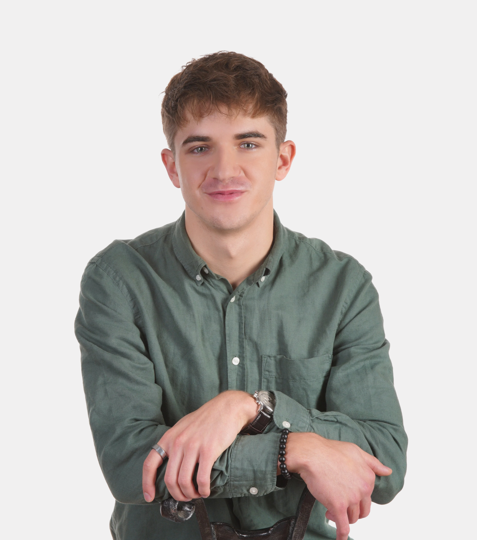 A young man with light brown hair and fair skin, wearing a green button-up shirt, smiles softly at the camera, with arms crossed and hands resting on a walking cane, against a plain white background.