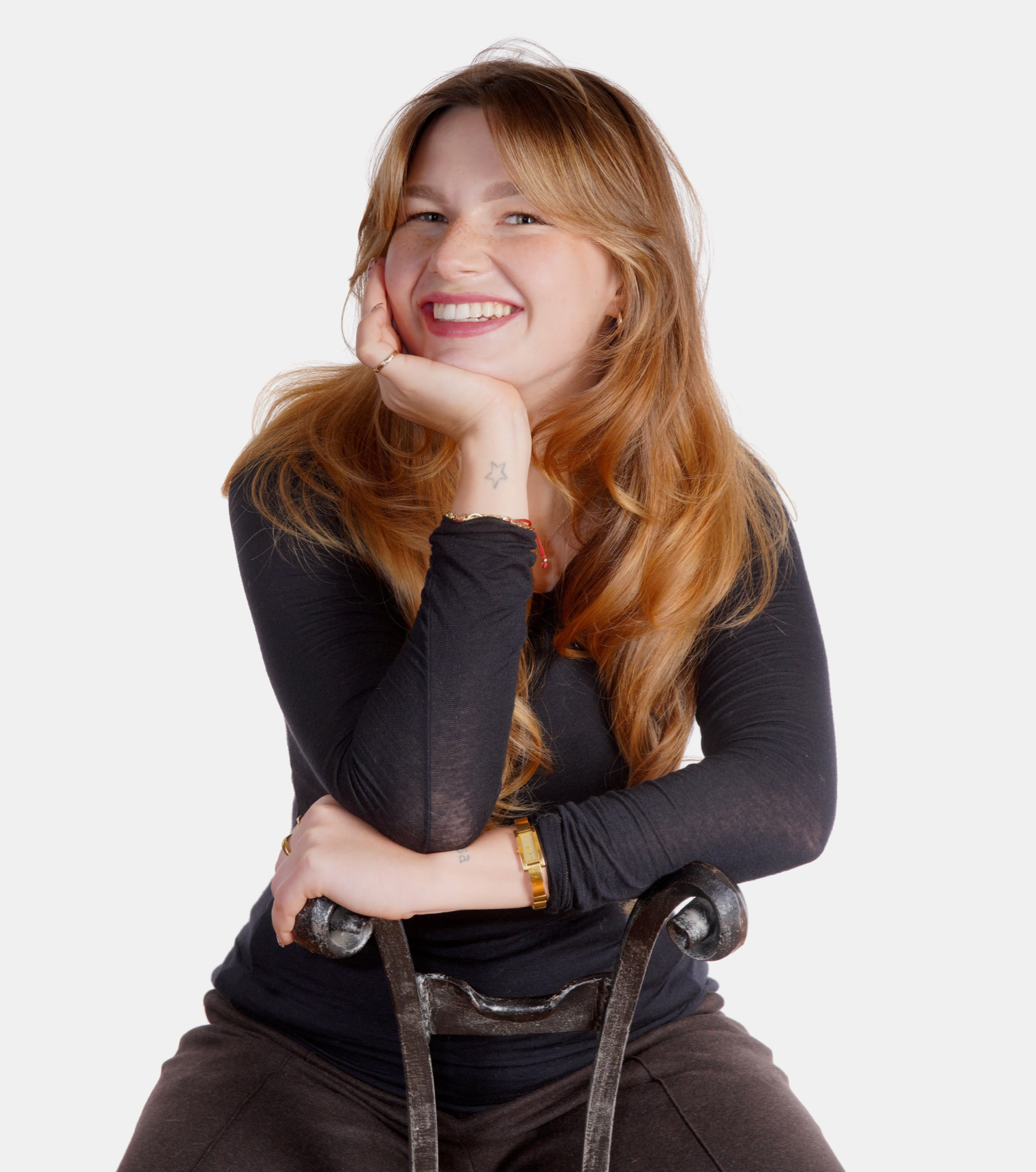 A young woman with red hair, wearing a black long-sleeve shirt, smiling and resting her chin on her hand, sitting in a relaxed pose on a chair, with a white background.