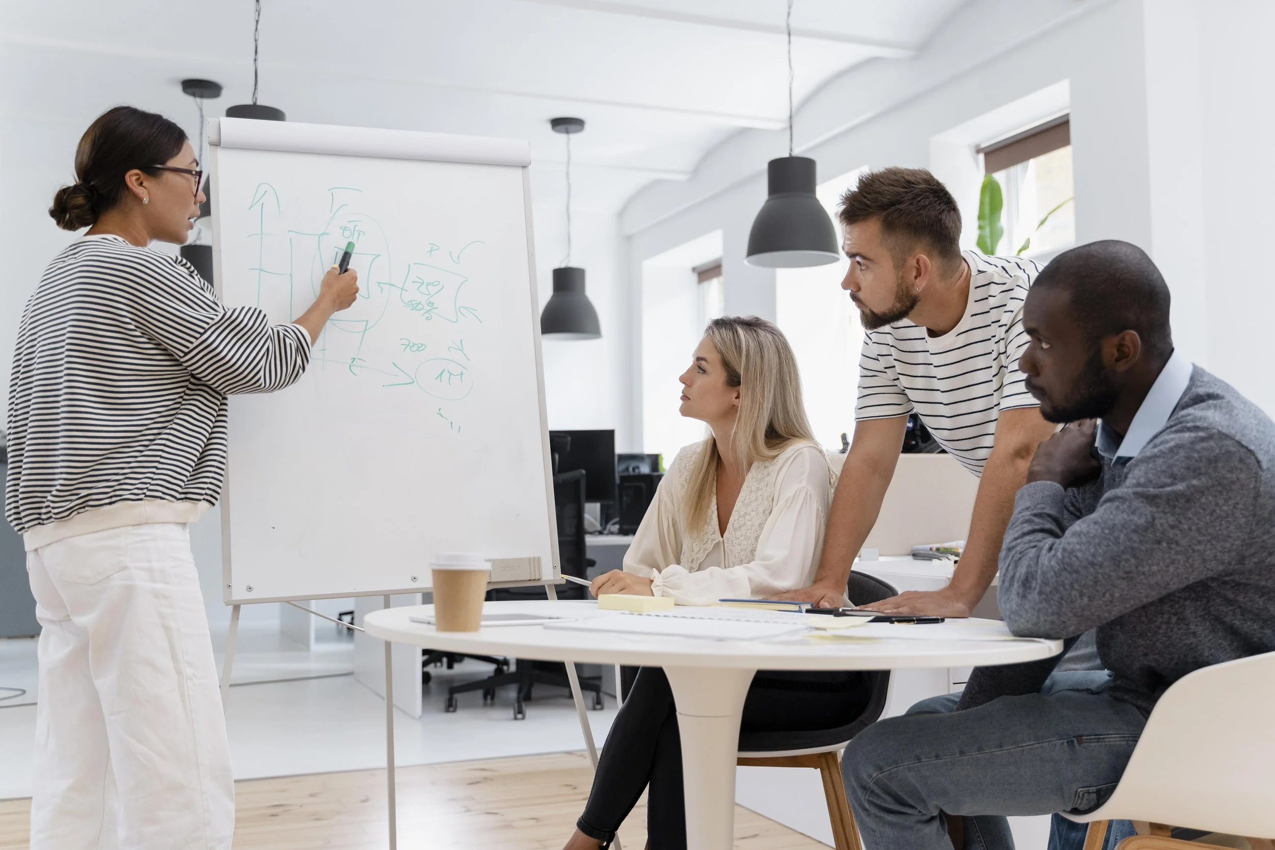 A woman in a striped shirt and white pants is writing on a whiteboard with diagrams, while three individuals observe: a blonde woman sitting at a white table, and two men, one with a beard and the other with dark skin, sitting and standing respectively, in a bright, modern office.