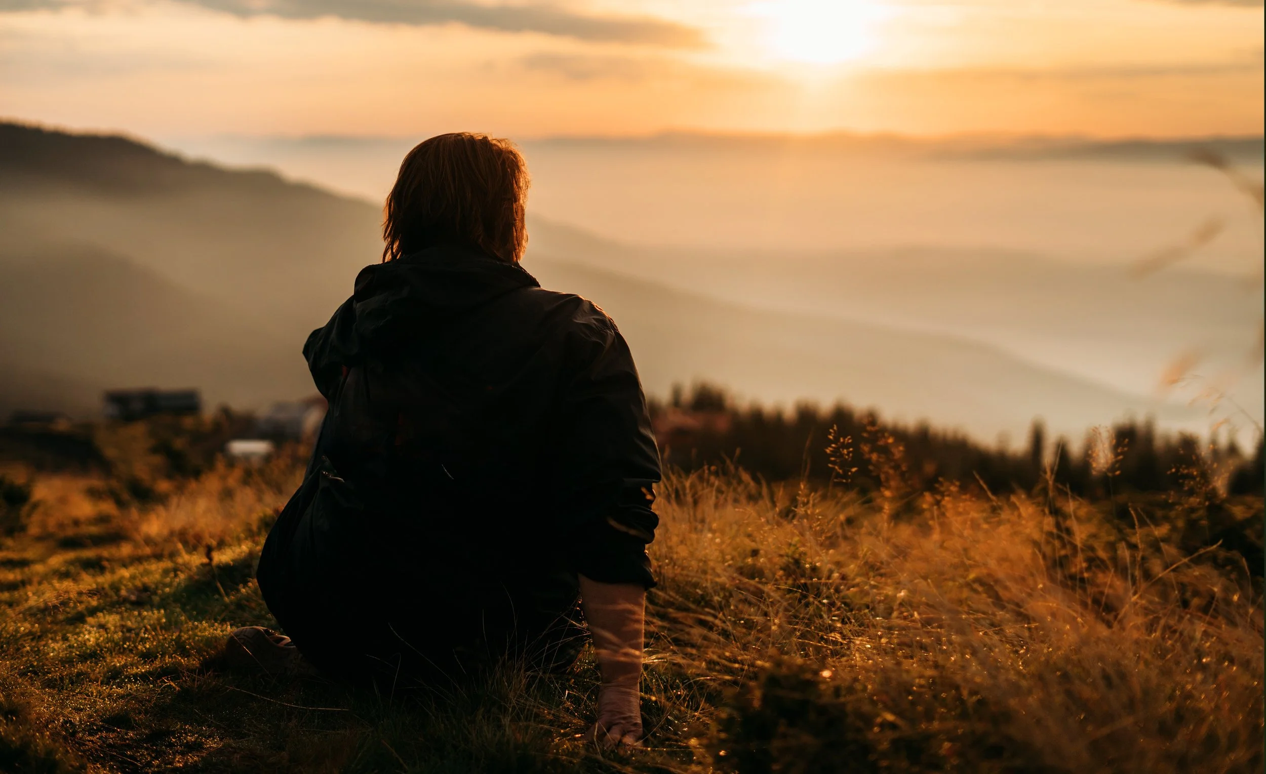 Άτομο καθισμένο σε ύπαιθρο, κοιτάζει τον ορίζοντα στο ηλιοβασίλεμα, συμβολίζοντας ηρεμία και αποκατάσταση.