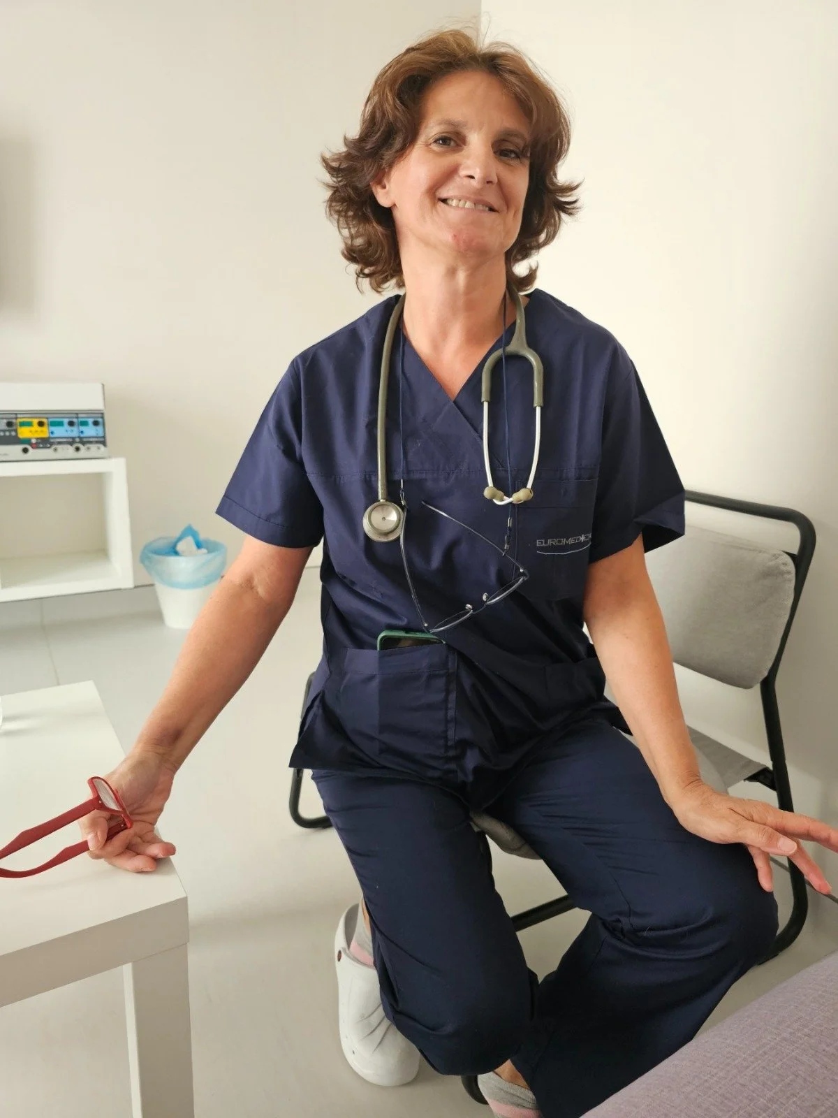 Ιατρός σε ιατρείο, με ιατρική στολή και στηθοσκόπιο, καθιστή και χαμογελαστή σε χώρο εξέτασης.