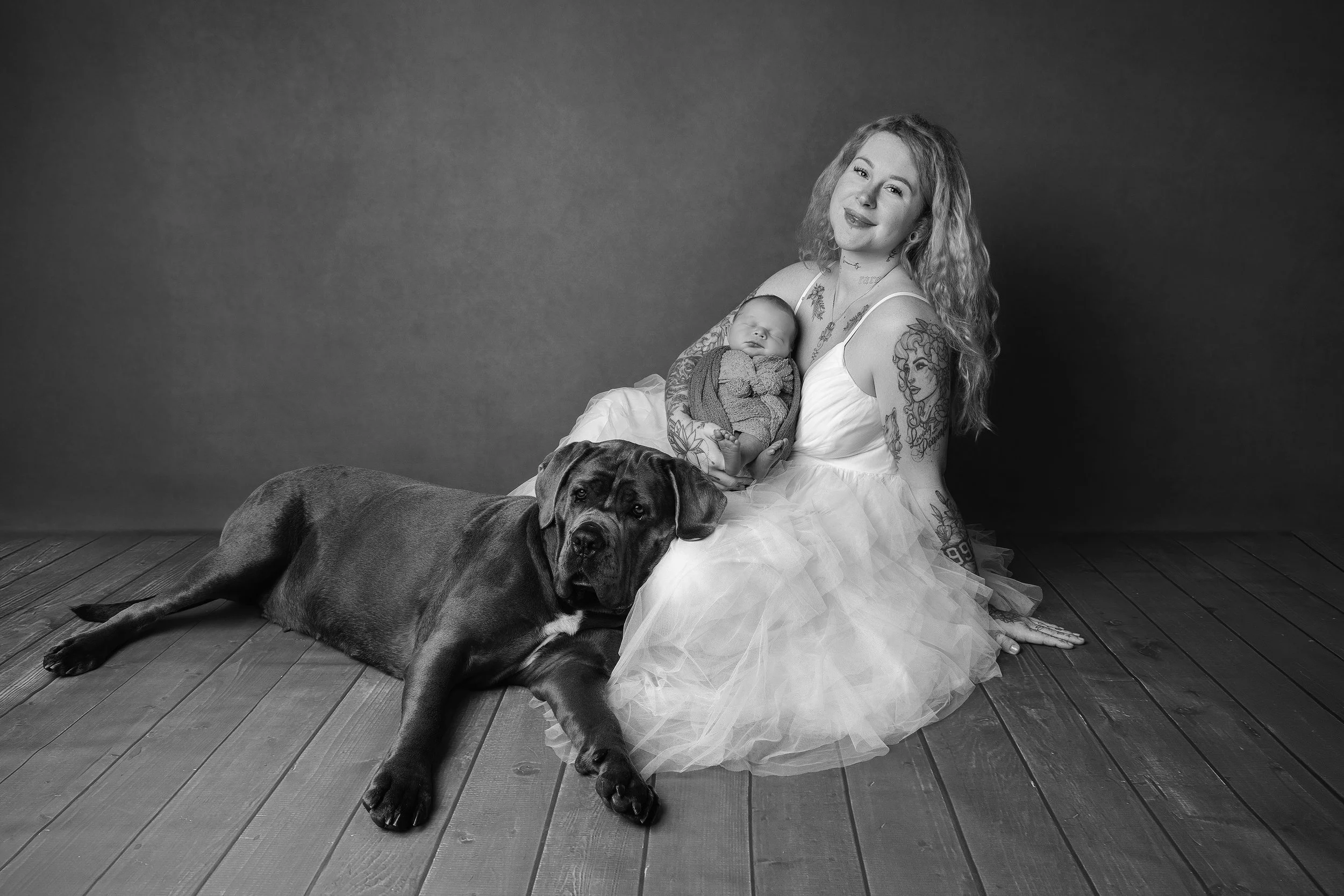 A woman with tattoos holding a sleeping baby, sitting on a wooden floor with a large dog lying beside her. The photo is in black and white.