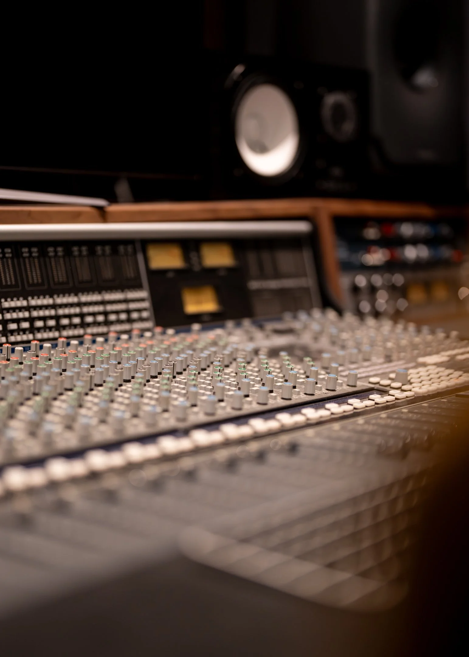 Close-up of a sound mixing console with various knobs and sliders in a recording studio, with studio speakers in the background.