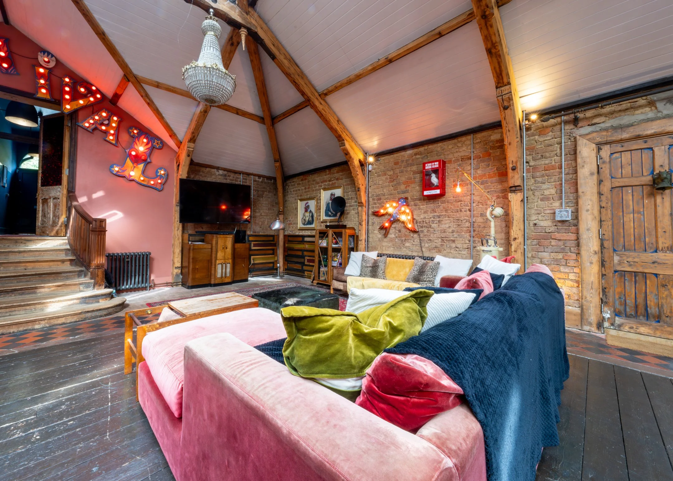 Interior of a living room with exposed brick walls, wooden beams, a pink velvet sofa with colorful pillows, a large flat-screen TV, wall art including a neon lion, a seagull, and a fire extinguisher, and eclectic decor with a chandelier and vintage f