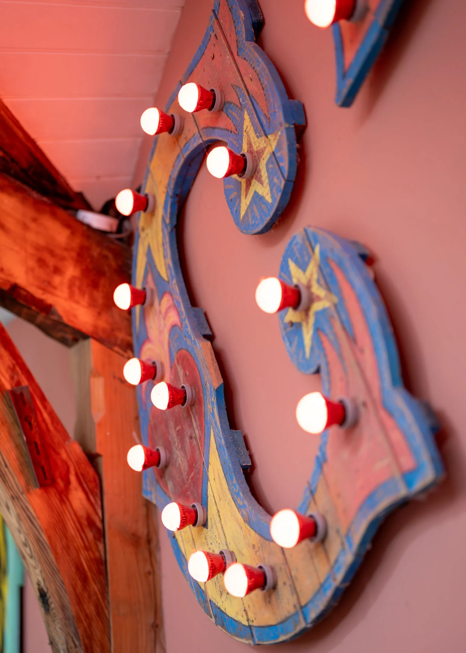 A decorative wooden sign in the shape of a heart with a colorful, vintage Americana design, featuring a star in the center and surrounded by small red and white light bulbs, mounted on a pink wall.