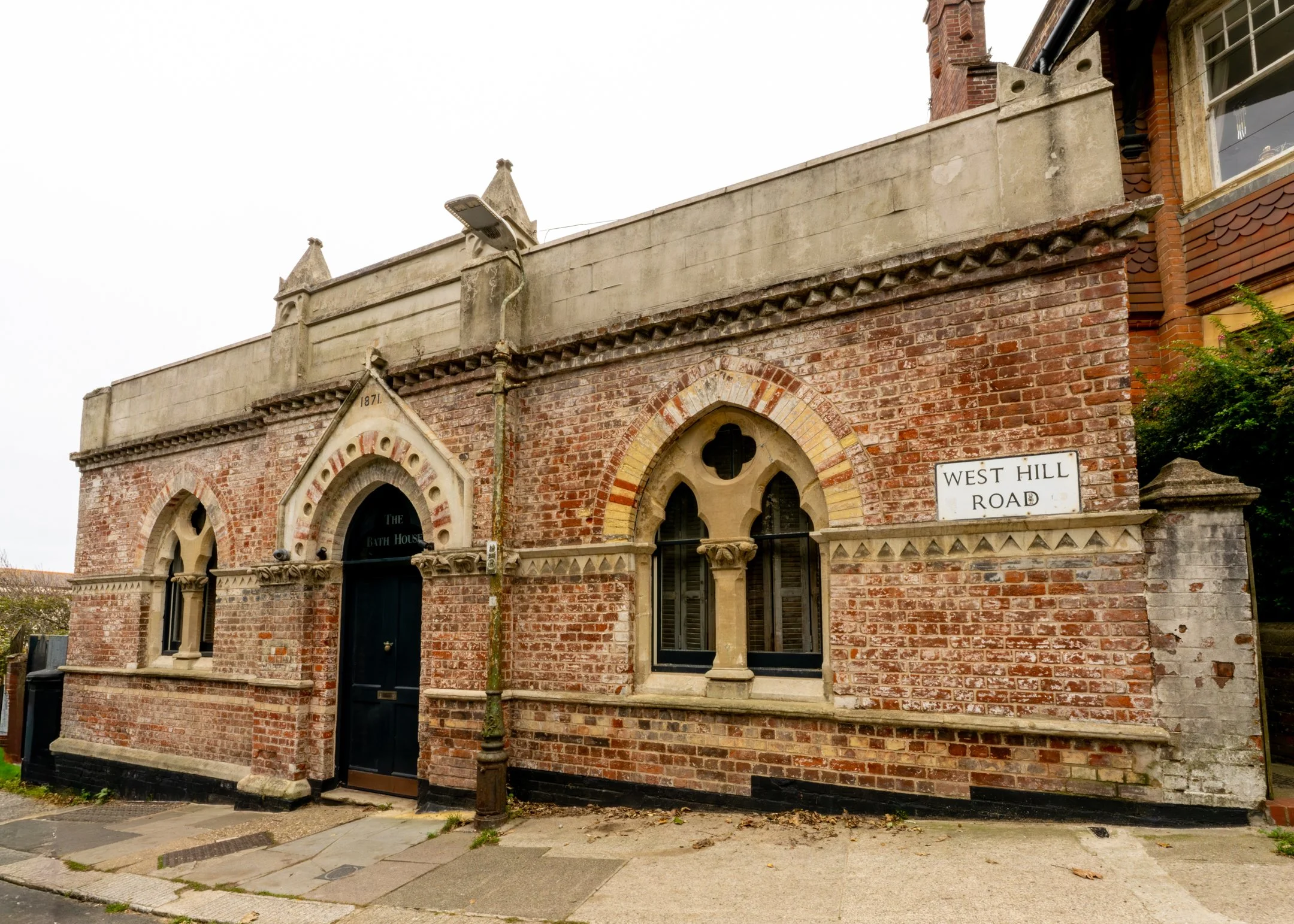 A small historic brick building with Gothic windows, a black door with a pointed arch, and a sign reading 'The Bath House'. There is a street sign that says 'West Hill Road'.