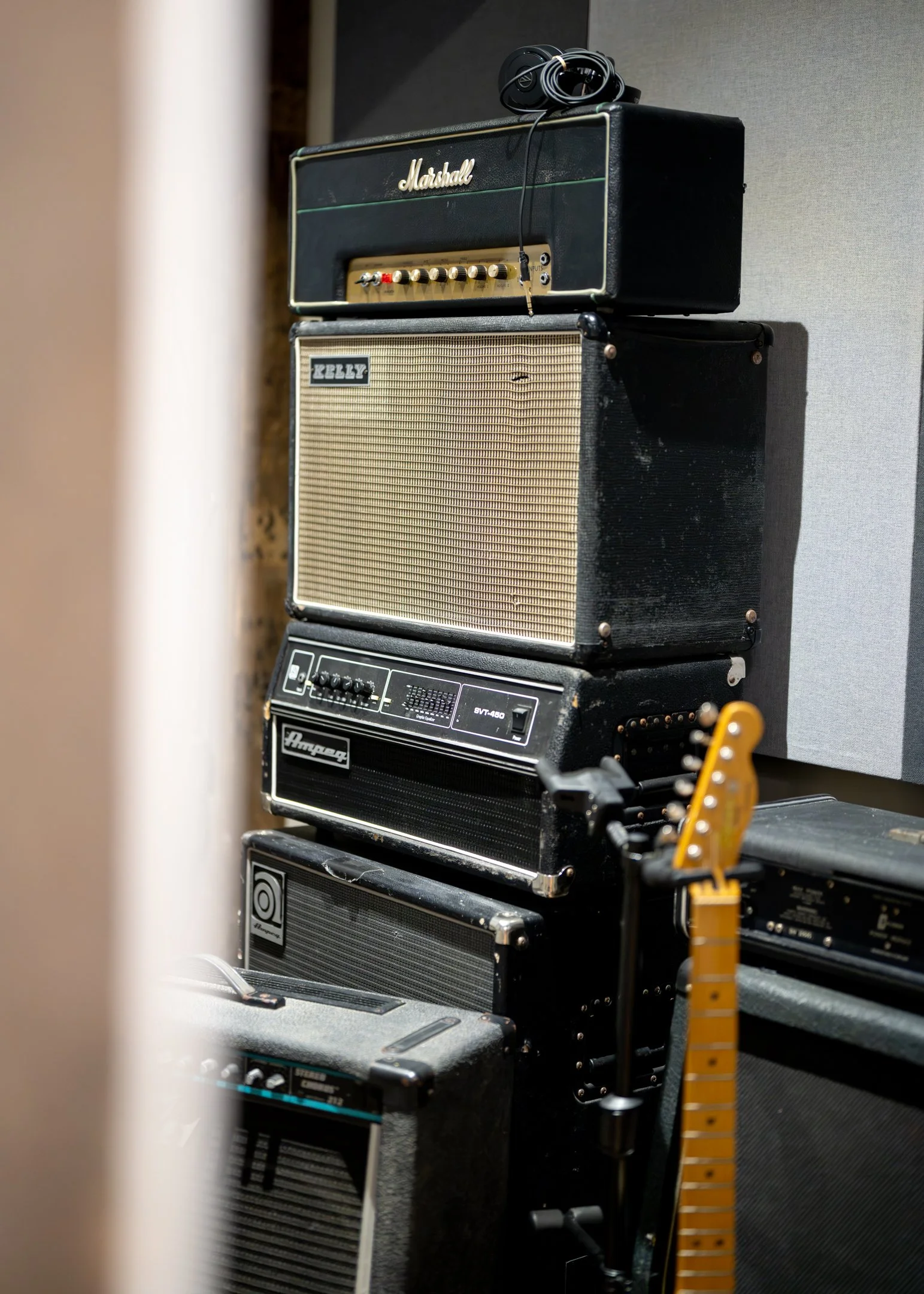 A stack of vintage guitar amplifiers with a guitar and an amplifier pedal in the foreground, placed against a wall.