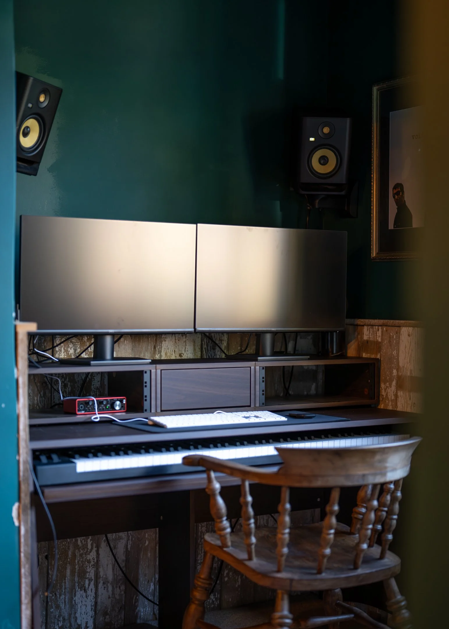 A music production studio setup with two computer monitors, a keyboard, a small audio interface, and two speakers mounted on the green wall, along with a framed picture of a man wearing sunglasses.
