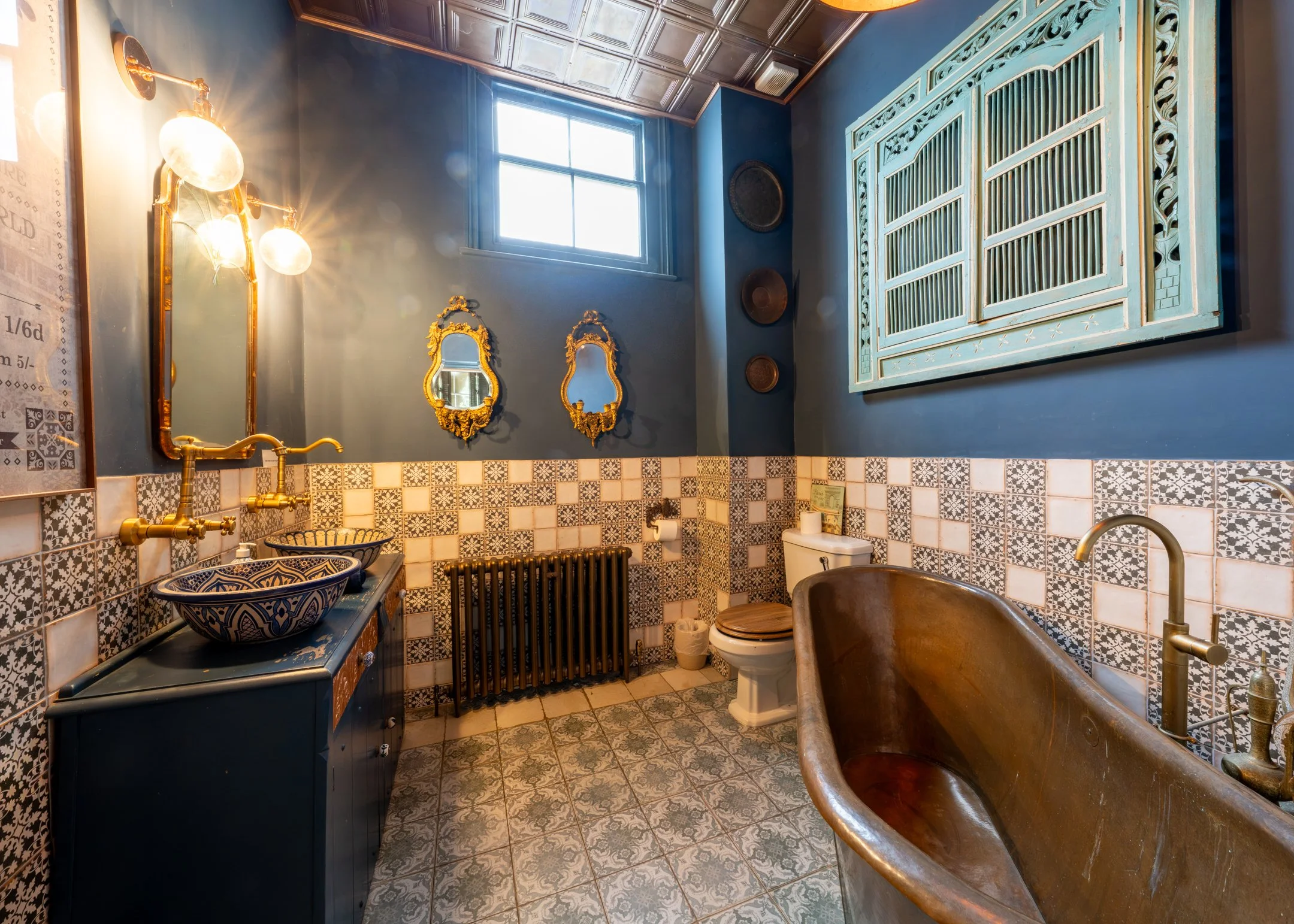 A vintage-style bathroom with a clawfoot bathtub, blue walls, patterned tiles, two small mirrors with ornate gold frames, a blue vanity with two vessel sinks, a toilet with a wooden seat, a brass fixture for the sink, decorative wall hangings, a wind