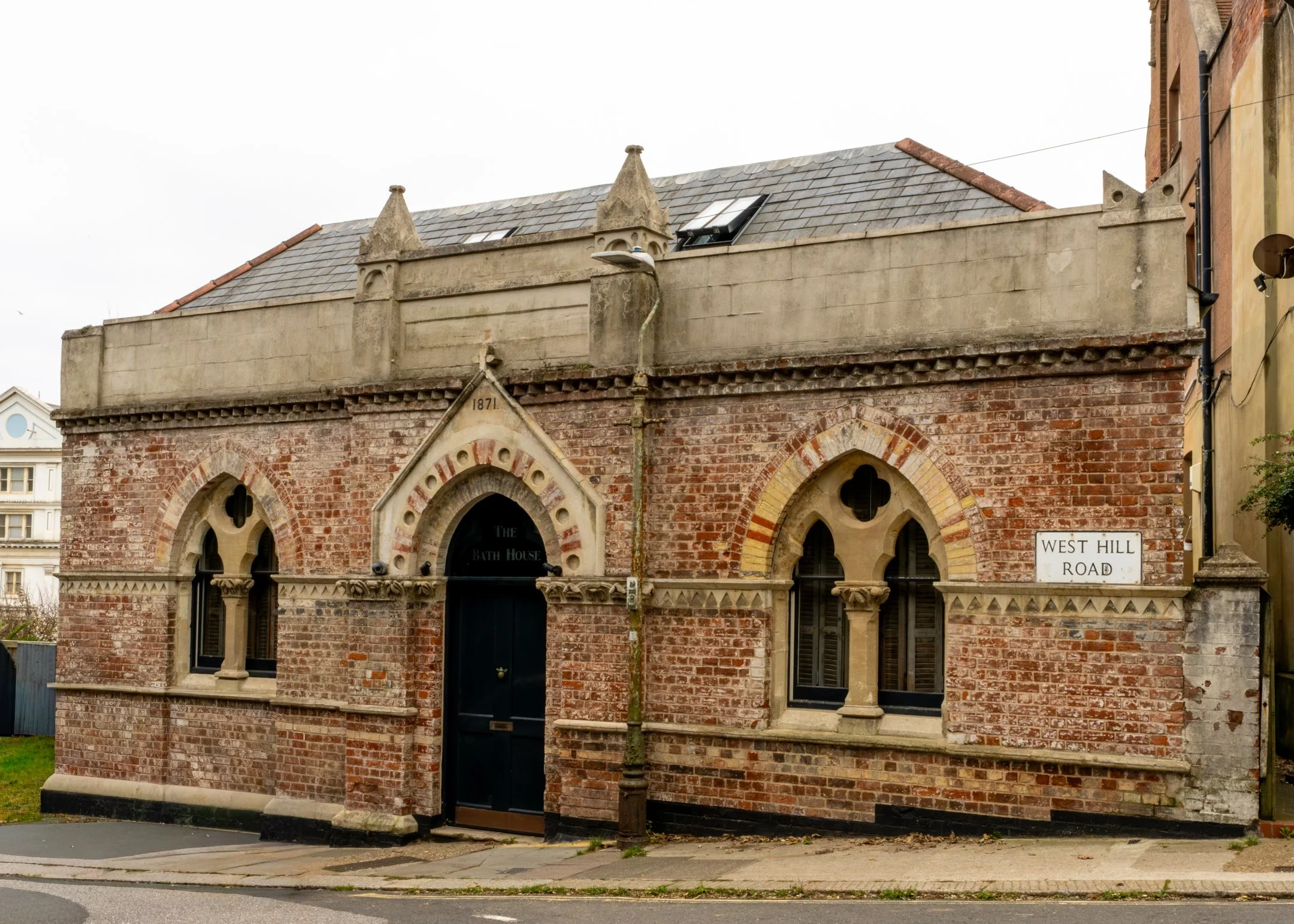 A small historic brick building with arched windows and door, labeled 'The Bath House,' with a sign on the side that reads 'West Hill Road,' street view with sidewalk and street curb.