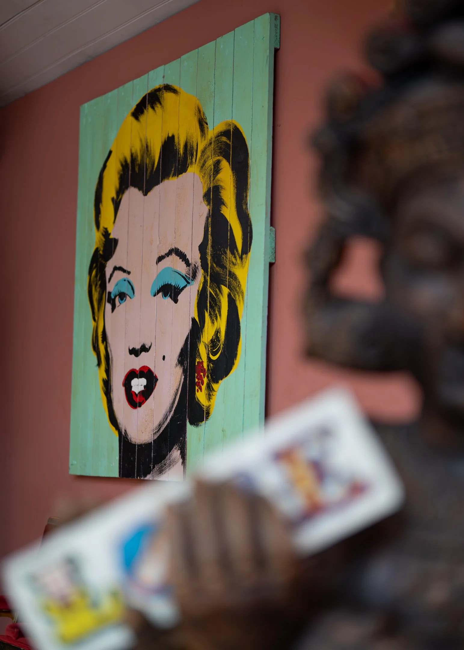 A colorful pop art portrait of Marilyn Monroe on a painted wooden panel, with a blurred foreground showing a person's hand holding a deck of playing cards and part of an intricate sculpture near the right edge.