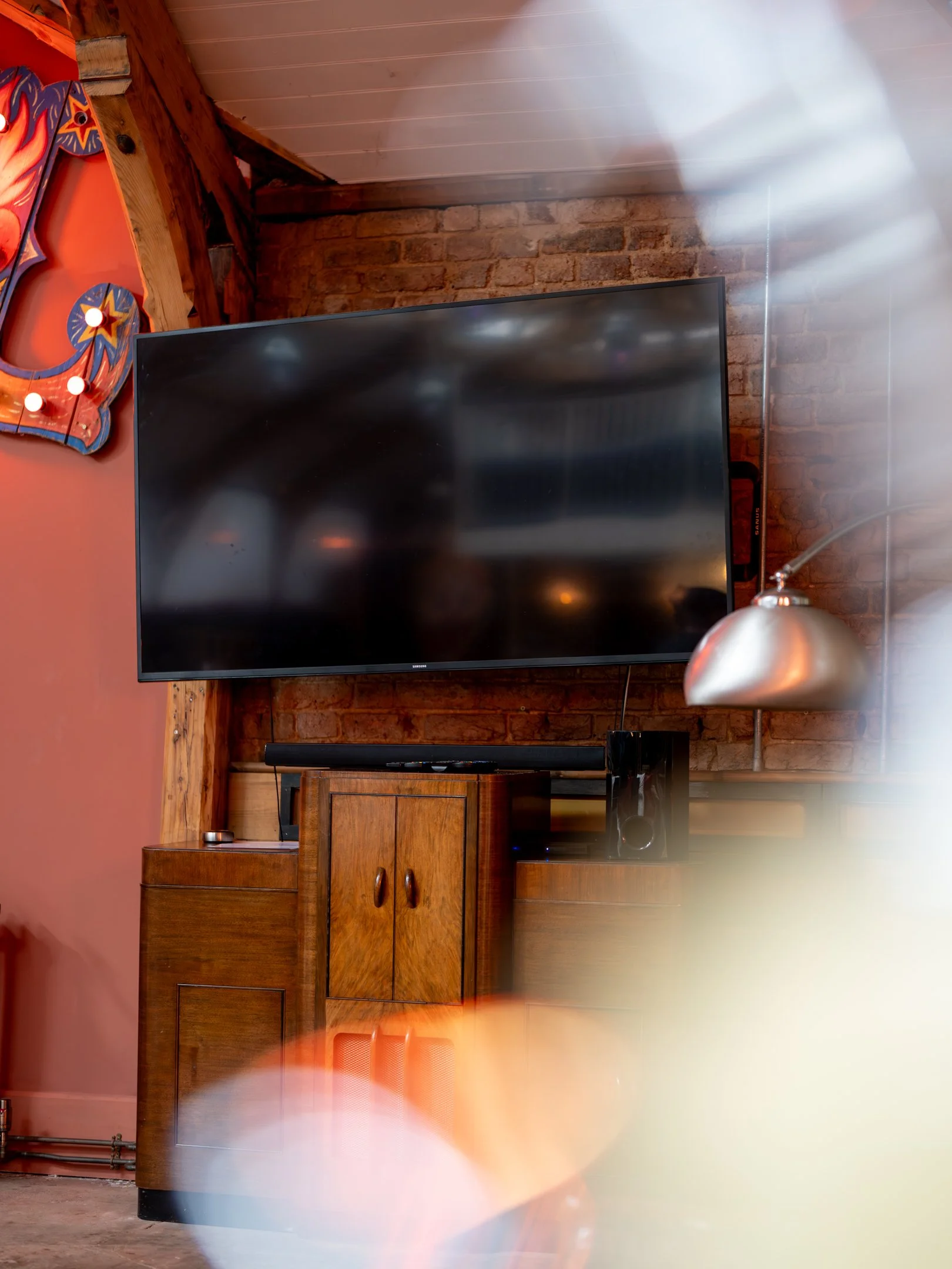 A flat screen TV mounted on a brick wall, with a wooden cabinet below it. To the right, there is a metallic hanging lamp. Part of a colorful decorative sign with lights and a wooden detail is visible on the left side of the image.