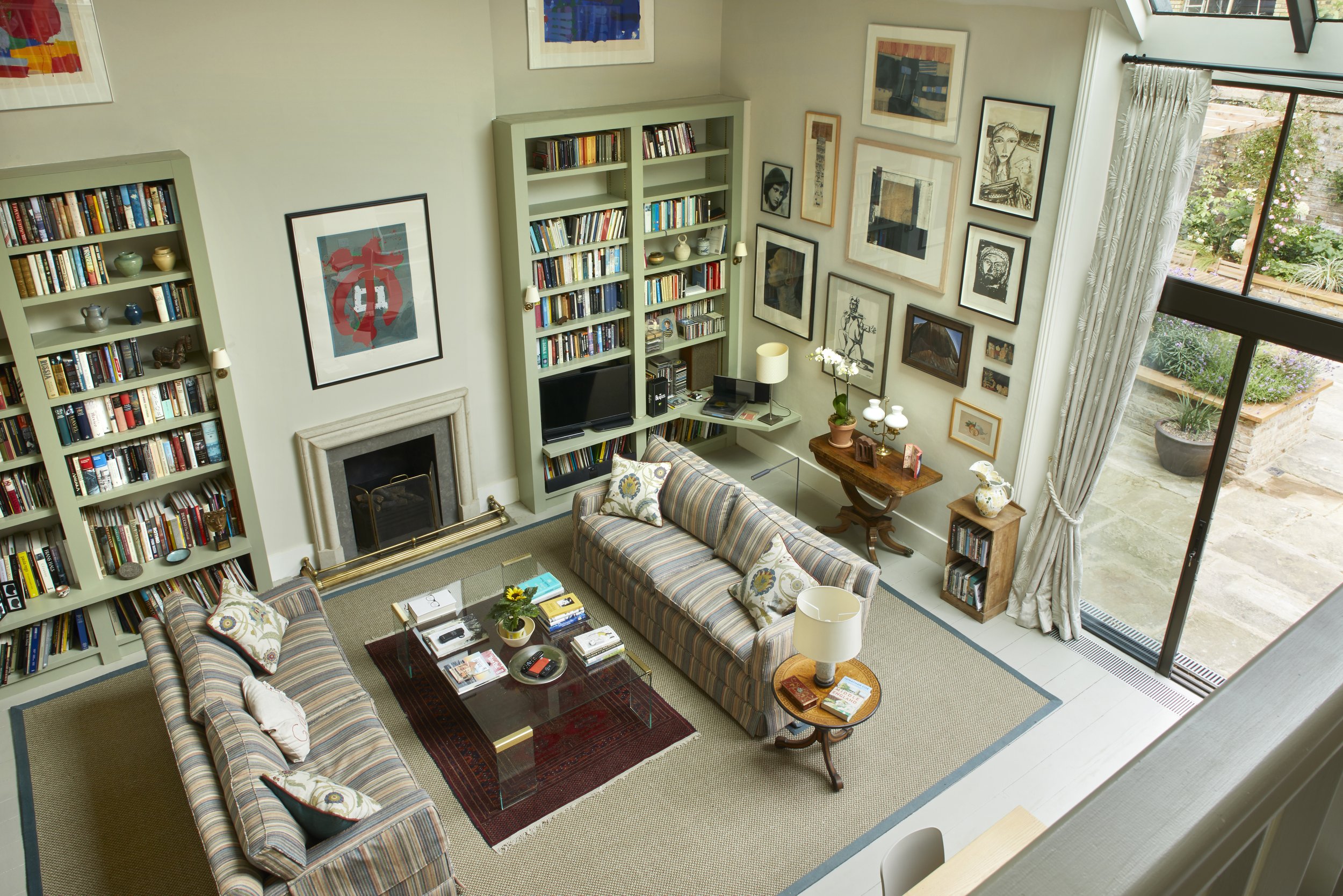 An aerial view of a cozy living room with two striped sofas, a glass coffee table with books and decor, a fireplace, multiple built-in bookshelves filled with books and decorative items, framed artwork on the walls, a computer desk with a monitor and