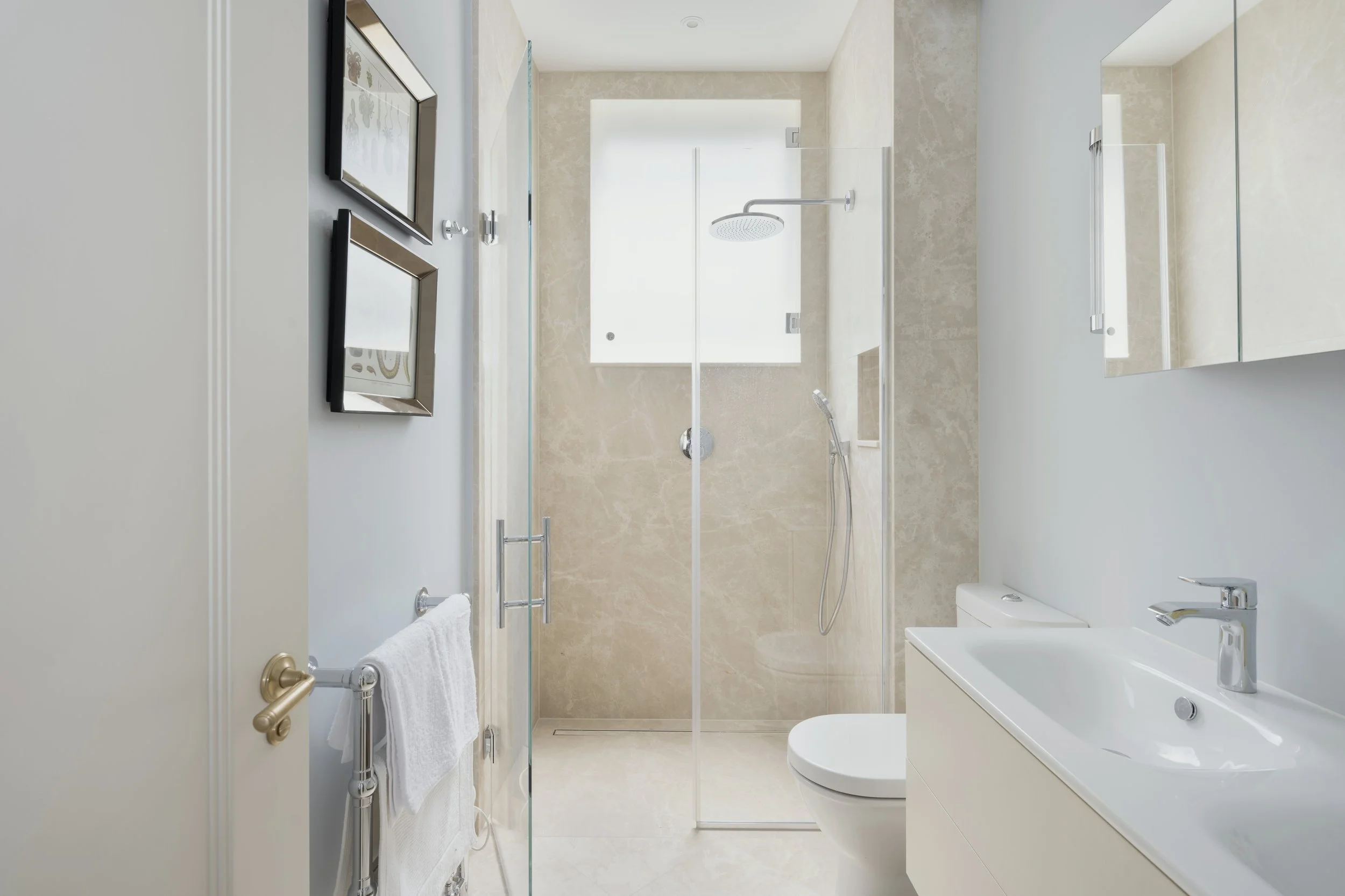 A modern bathroom with a walk-in shower, a white toilet, a white vanity with a sink, and a mirror. The shower has a rainfall showerhead and a handheld shower. There are two framed pictures on the wall, a towel rail with a white towel, and a large win