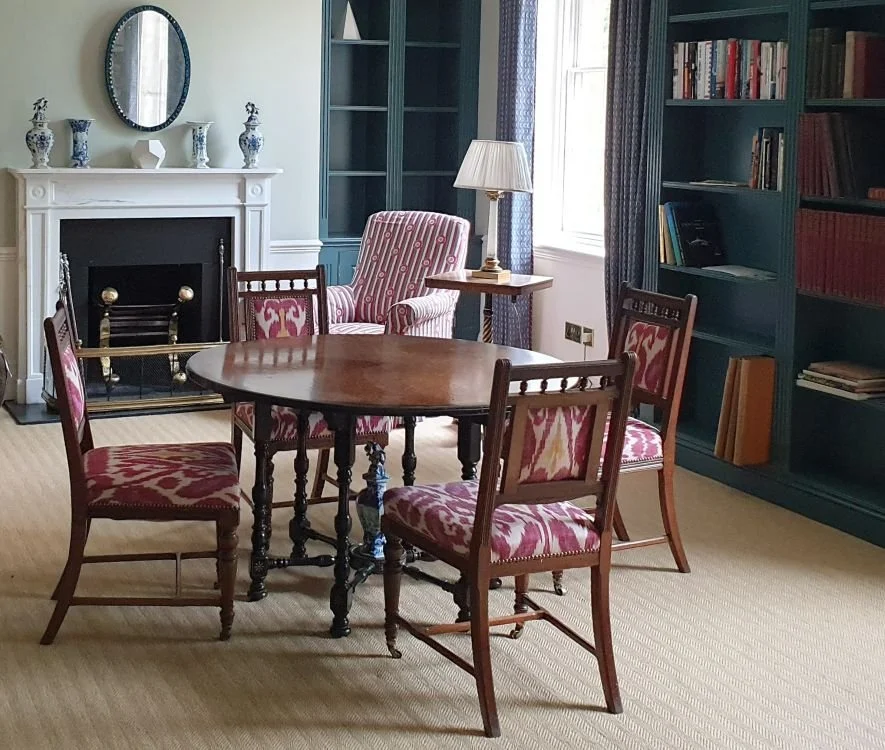 A decorated sitting room with a round wooden table, four wooden chairs with red and white patterned upholstery, an armchair with matching fabric, a bookshelf, a fireplace with decorative vases, and a lamp beside the armchair.