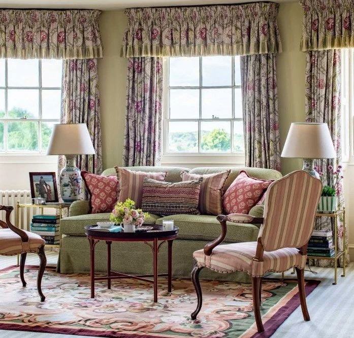 Living room with a green sofa, striped accent pillows, and a floral armchair. Two large windows with floral curtains, two table lamps, a floral centerpiece, and framed photograph on a side table.