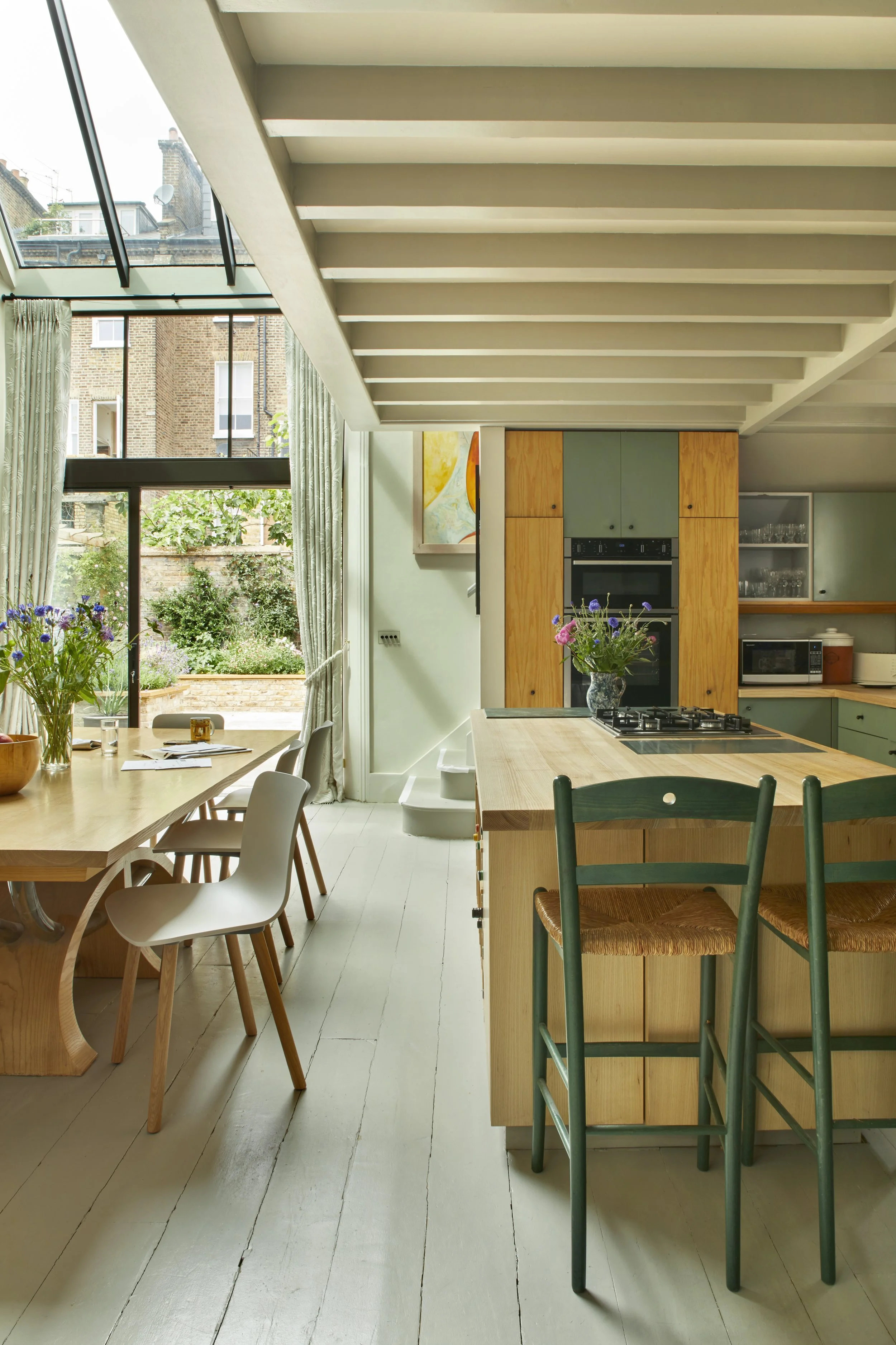 Bright kitchen and dining area with large window, wooden table with chairs, kitchen island with stools, and plants.