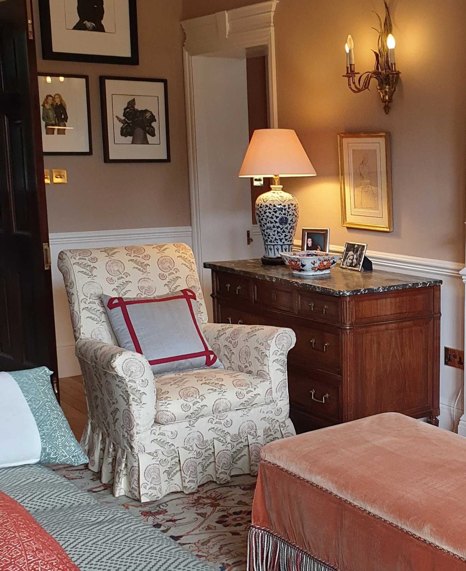 A cozy living room corner with a patterned armchair, a wooden sideboard with a marble top, a decorated lamp, framed photographs, and wall art.