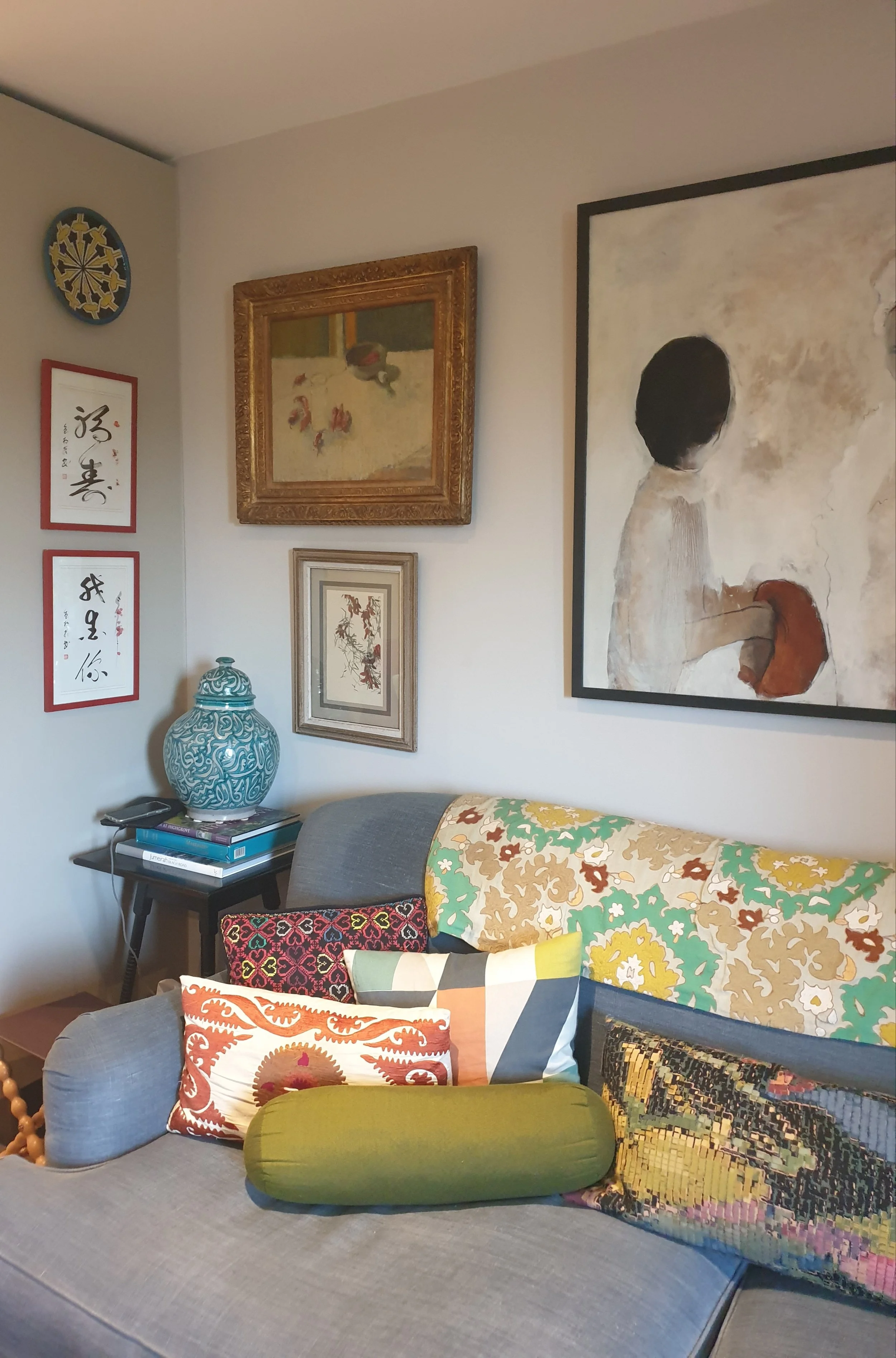 A living room corner with a gray sofa decorated with colorful patterned pillows, wall art, and a blue decorative vase on a side table.