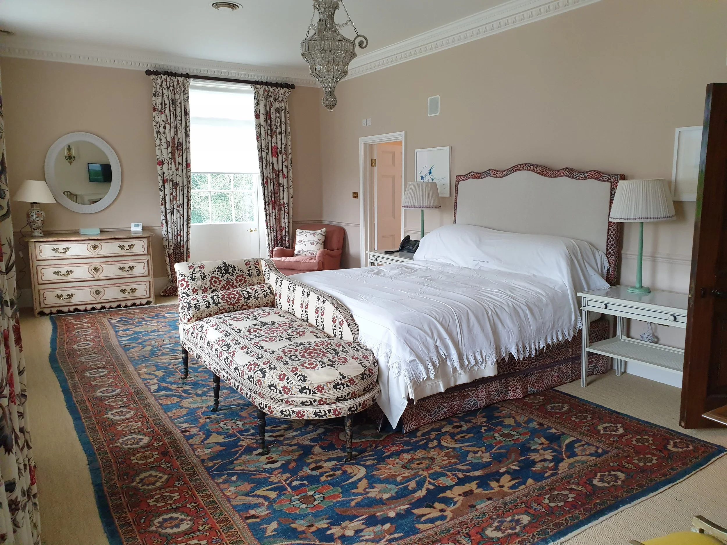 A luxurious bedroom with a large bed, adorned in white linens and surrounded by vintage-style furniture, including a matching upholstered bench at the foot, side tables with lamps, and an armchair near a window with floral curtains. The room features