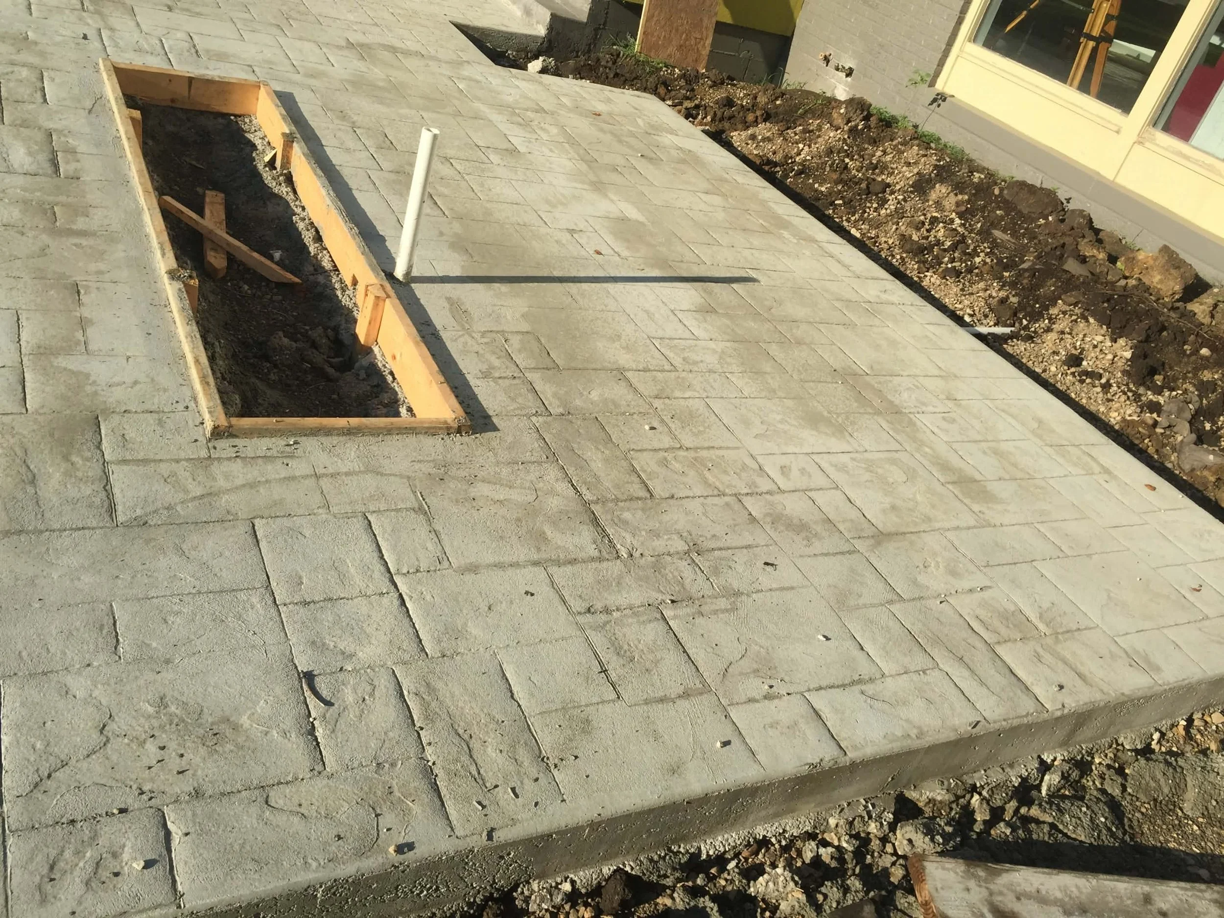 Ongoing outdoor construction of a paved walkway with a rectangular hole and a white pipe sticking out, surrounded by soil and dirt, with custom rectangle fire pit opening