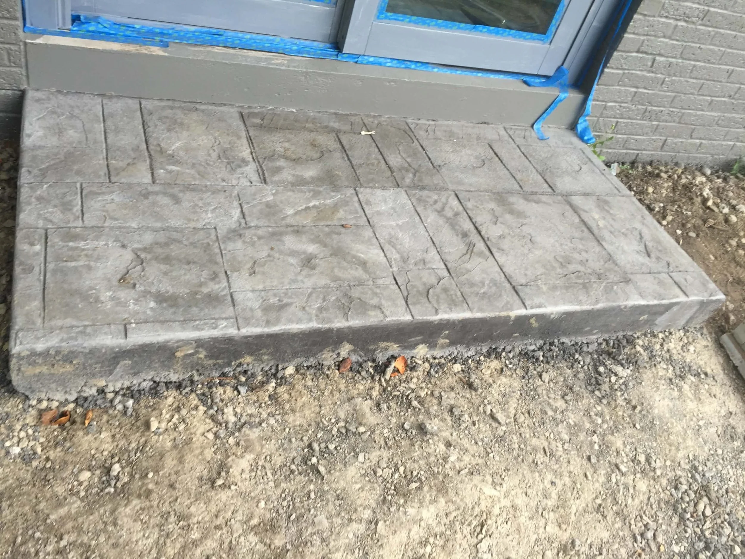 Concrete porch with patterned paving stones in front of a door, with blue painter's tape around the door frame, and a dirt area surrounding the porch.