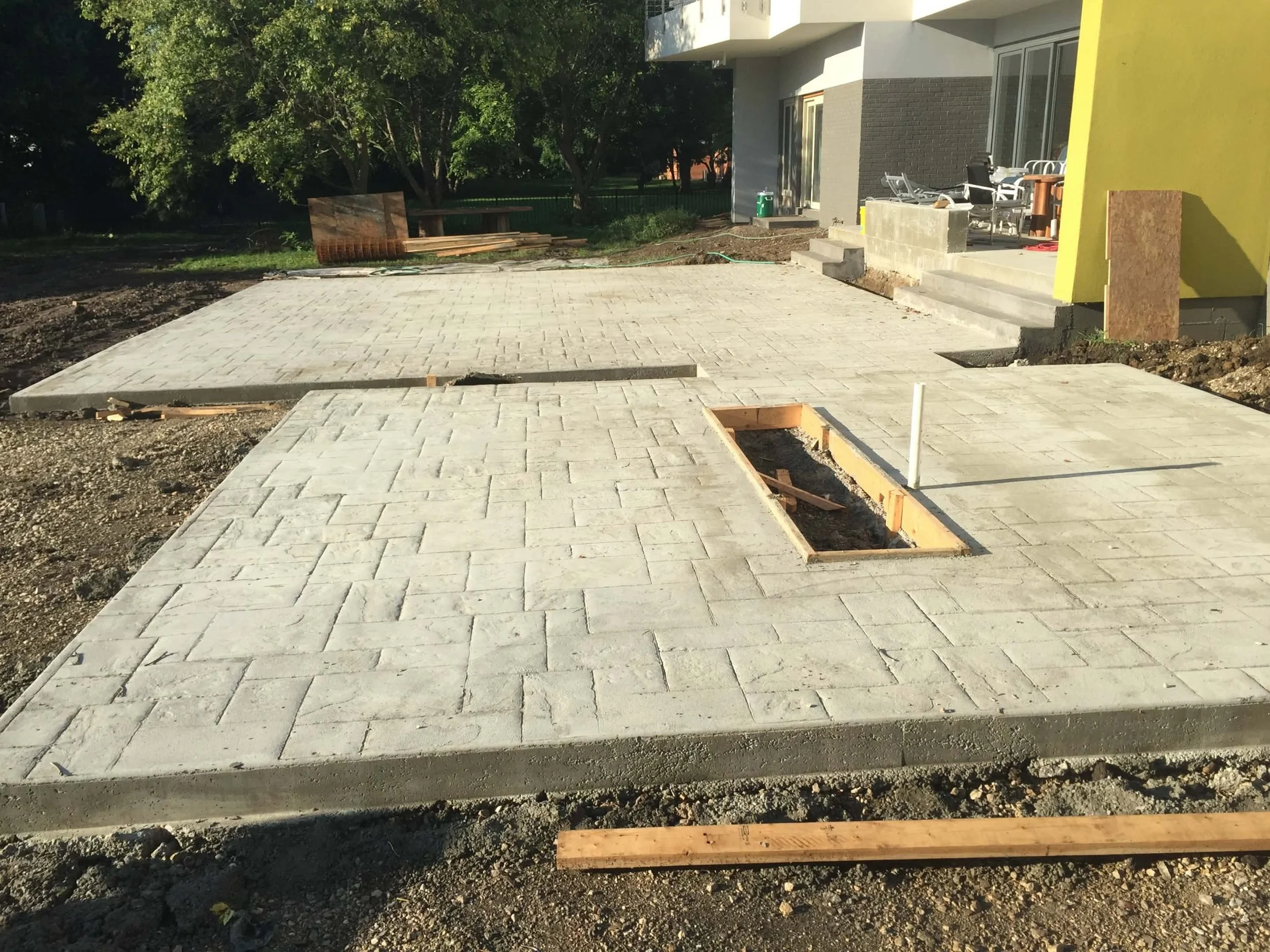 Construction site with a newly laid concrete paver patio, wooden forms for future installation, and stairs leading to a modern building with outdoor furniture, surrounded by dirt and construction materials in preparation for a custom fire pit
