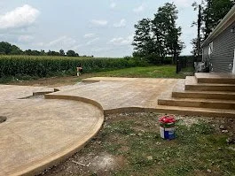 A backyard with a newly poured custom stamped concrete patio, partially finished with stairs leading up to a house, and a bucket of paint or stain nearby by Rooks Construction and concrete.