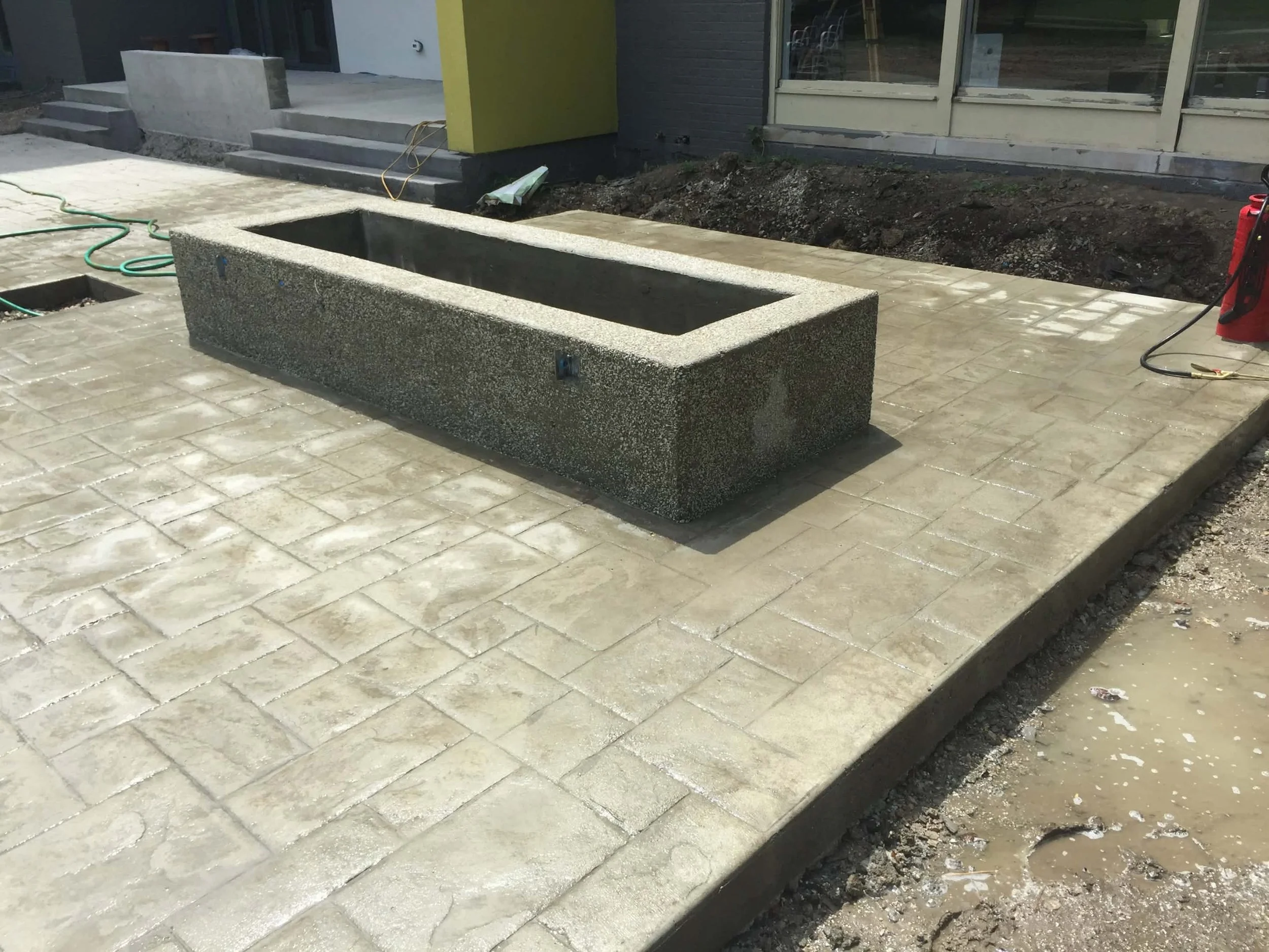Construction site with a rectangular concrete fire pit on paved ground, and a building with glass doors in the background
