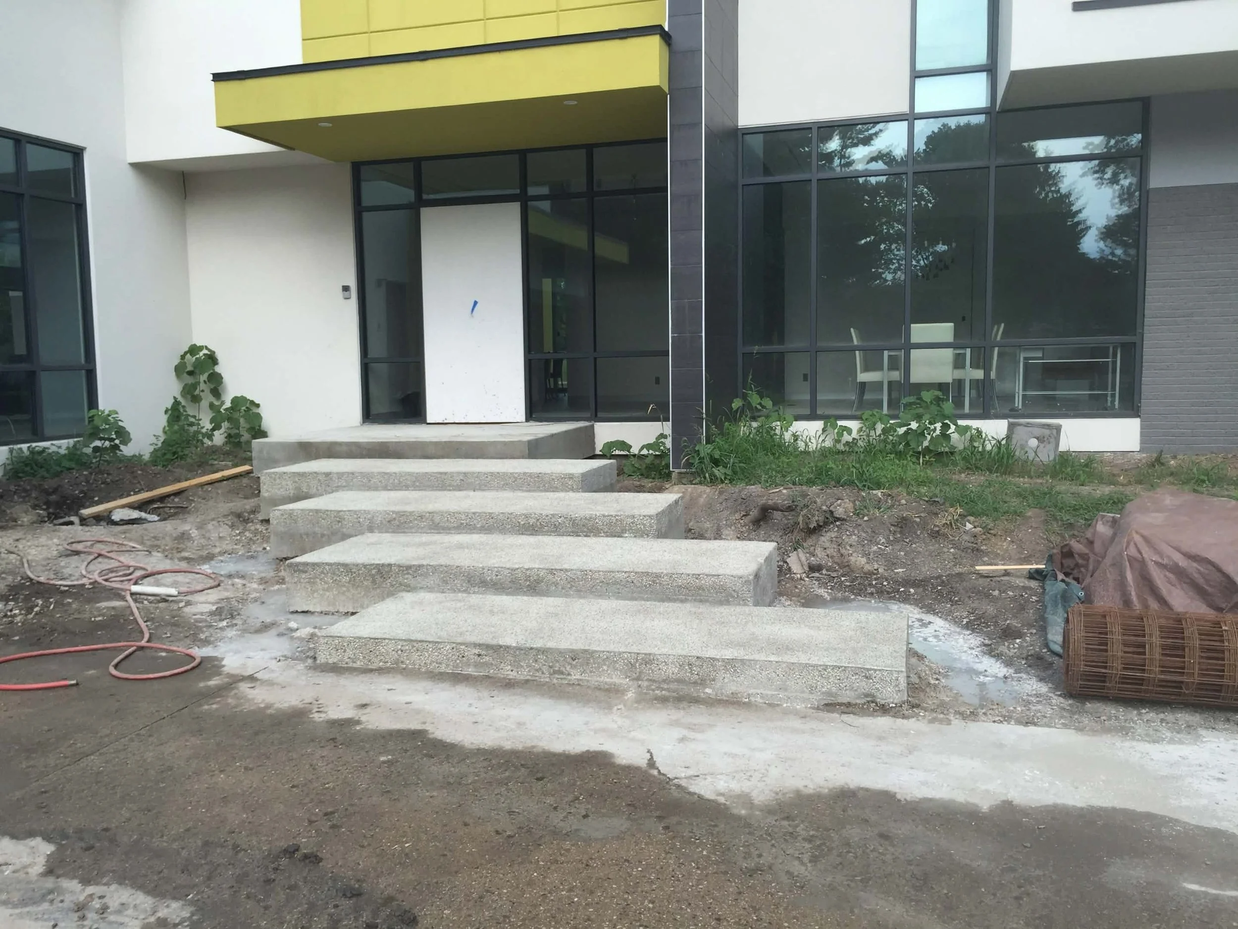 Construction site of a modern house with concrete stairs leading to a white door, large glass windows, and surrounding dirt and construction materials.